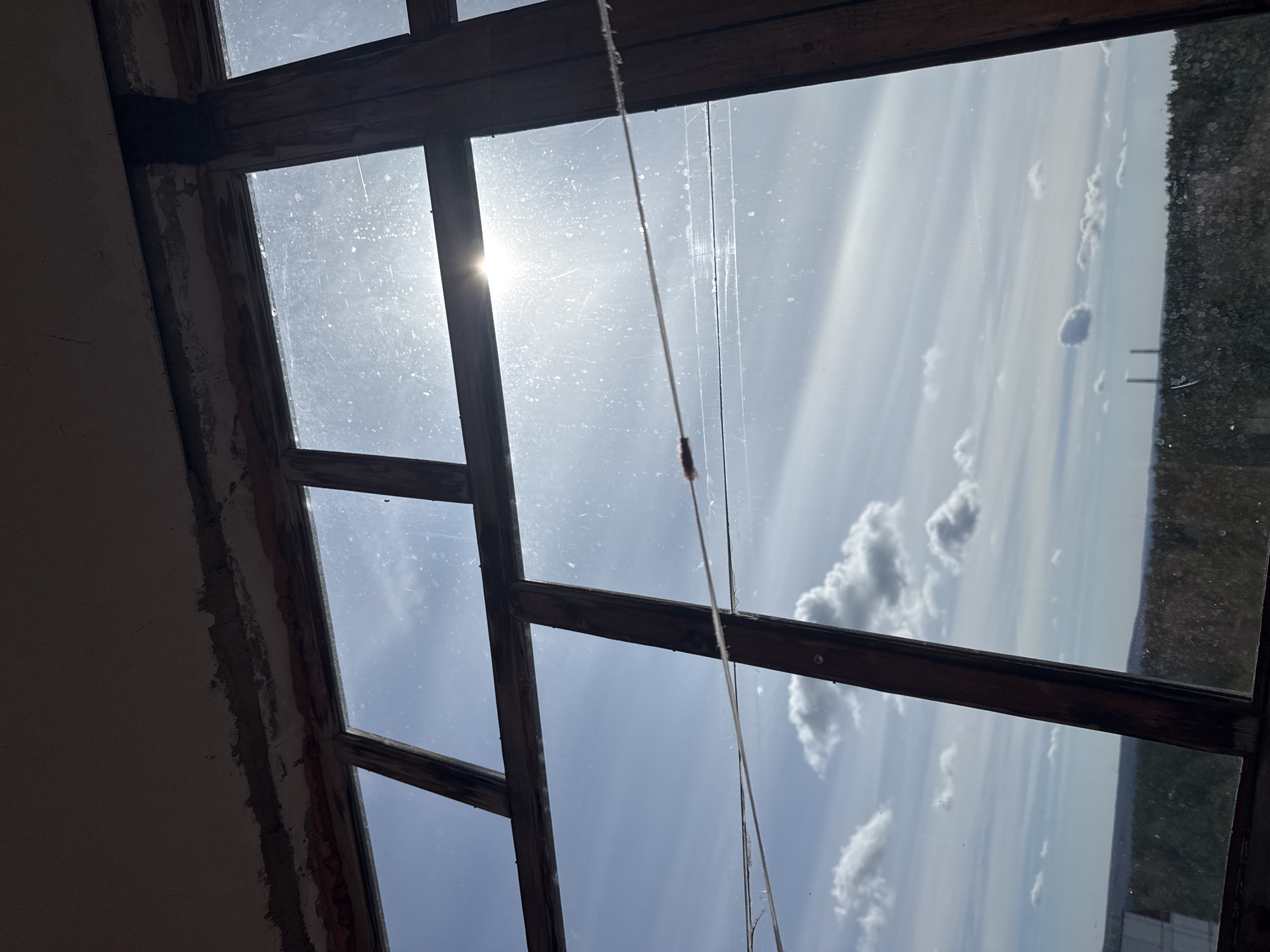 The image presents an upward-angled view through a multi-pane window, revealing a bright sky and distant landscape from an interior in Chaikovsky, Russia. The window features dark, weathered wooden frames, suggesting an older structure or a space undergoing renovation. On the left side, an unfinished wall with exposed plaster or concrete adds to this impression.
The glass panes are visibly dirty and dusty, covered in numerous spots and streaks, particularly noticeable where direct sunlight pierces through. A brilliant sun flare is prominently featured in the upper left section, radiating intense light. The sky outside is a clear, light blue, sparsely dotted with fluffy white cumulus clouds. Towards the far right, a dark, textured area hints at distant trees or dense vegetation.
A thin, light-colored string or rope descends vertically from the top window frame. A small, dark, elongated object is attached to its lower part, possibly an insect cocoon, a small weight, or part of a window mechanism. The strong illumination confirms it is daytime, likely morning or afternoon. No human activity or visible text is present in the frame. The scene conveys a quiet, observational moment through an aged, imperfect window.