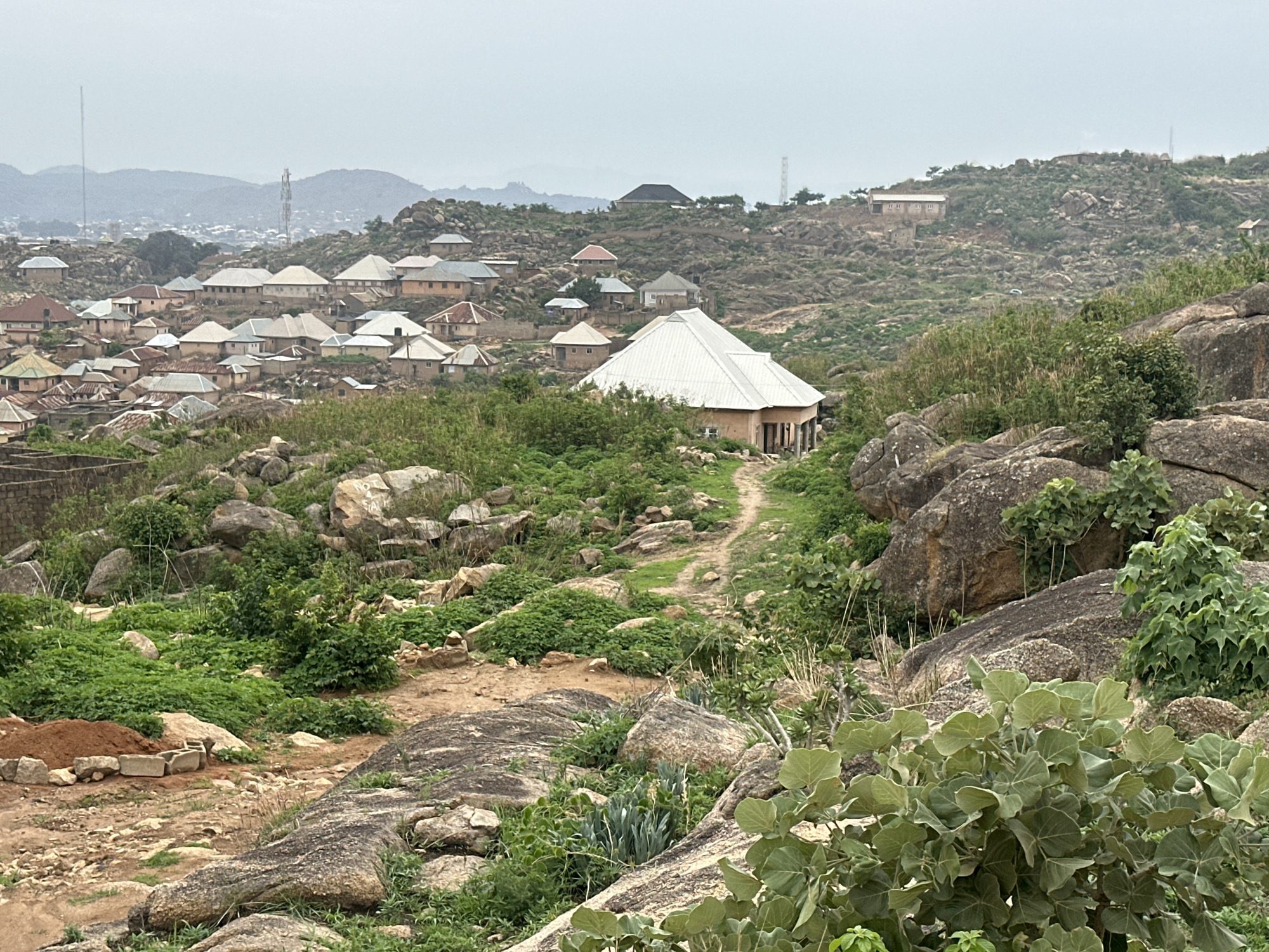 The image depicts a hilly landscape in Jos, Nigeria, dotted with houses and characterized by rocky terrain interspersed with vegetation. A cluster of houses with diverse roof colors is situated on the left side of the frame. A prominent single-story house with a large, white, pyramid-shaped roof is positioned on a slope in the mid-ground. The foreground showcases boulders and shrubs, and a dirt path leads up towards the house with the white roof. In the background, more houses are scattered across the hills, with utility poles visible in the distance. The sky is overcast.