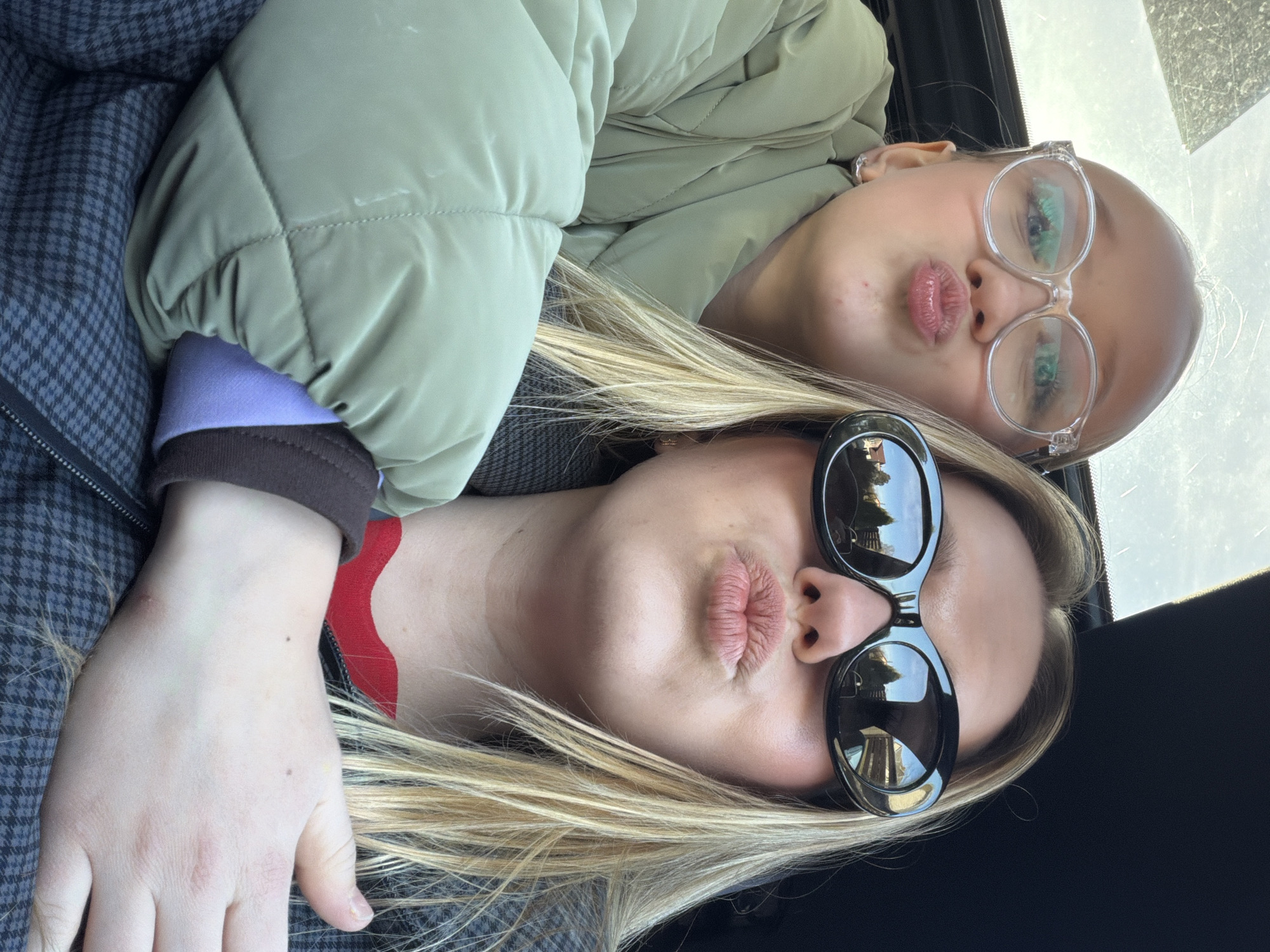 The image shows two people posing cheek-to-cheek, likely in a car. The individual in the foreground has blonde hair and is wearing sunglasses with dark lenses. Their lips are pursed into a kiss. The other individual, positioned above and to the side, is wearing clear glasses and also pursing their lips. The person in the foreground is wearing a red and black top with a grey jacket, whilst the other is wearing a green puffer jacket. The image appears to be taken from within a car, possibly in Dreiliņi, Latvia, due to the reflection in the sunglasses. The reflections in the sunglasses show what may be buildings.