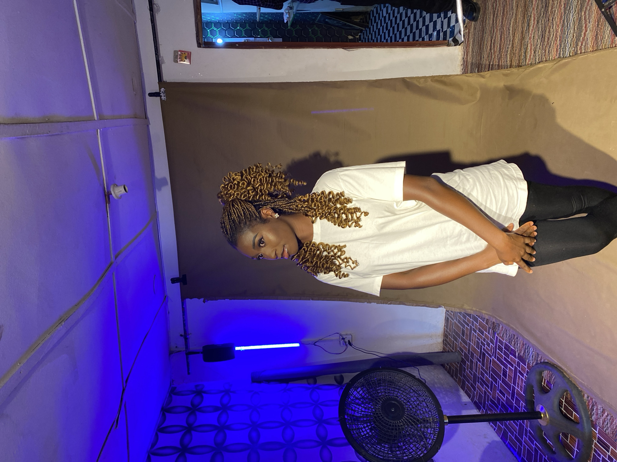 A young, dark-skinned woman with braided hair stands against a brown backdrop, possibly in a studio setting. She wears a white T-shirt and black leggings. The room is lit with blue light on one side, revealing patterned tiles on the floor. A black fan stands in the foreground. A partial view through a doorway reveals a room with checkered flooring. The location appears to be Vom, Nigeria.