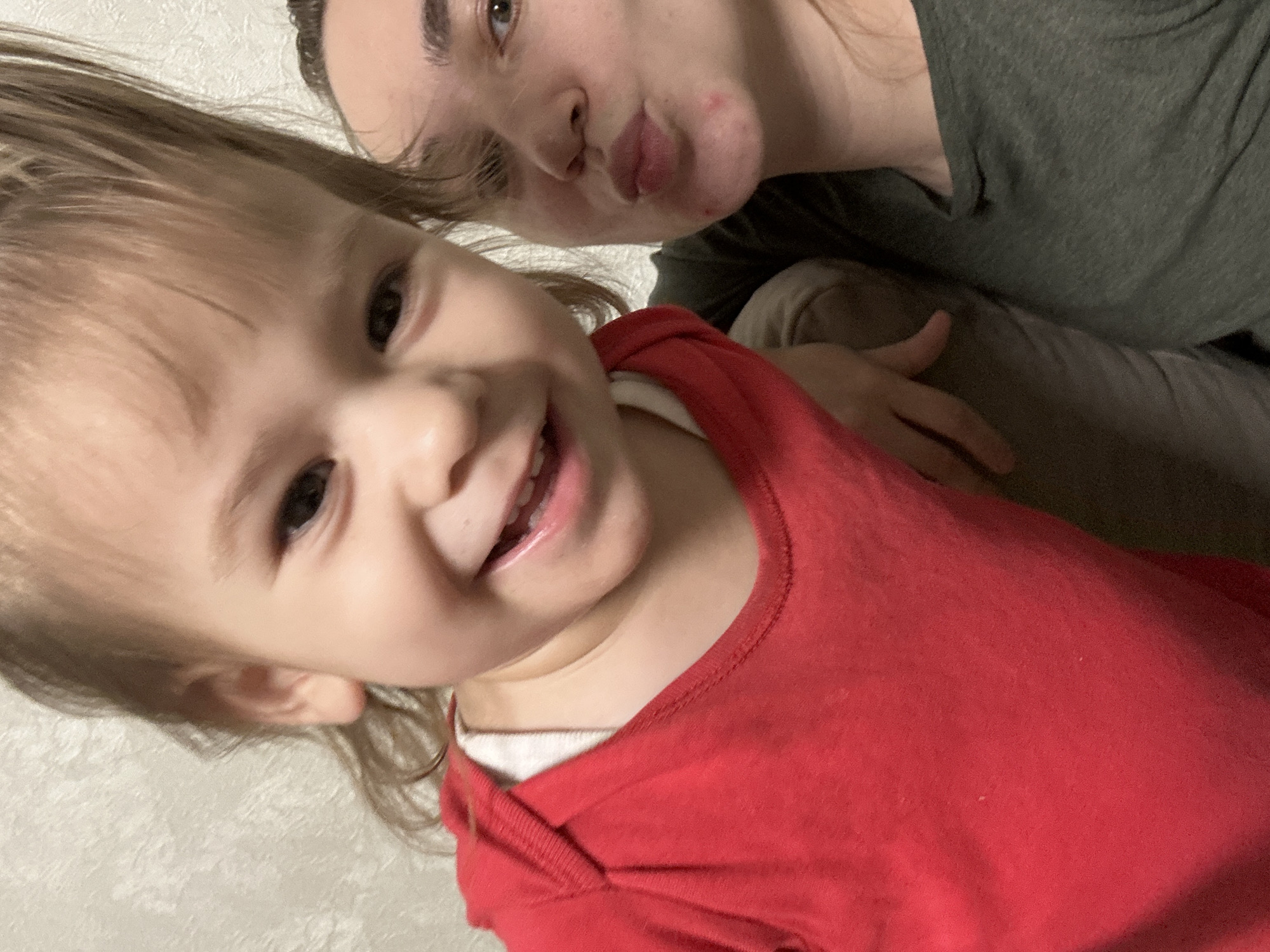 A selfie taken indoors shows a young child and an adult. The child, in the foreground, is wearing a red shirt with a white underlayer and is smiling, revealing teeth. The adult, partially visible in the background, is wearing a dark green shirt. The environment appears to be a room with a textured, light-colored wall in the background. The location is given as Uva, Russia, but there is no contextual evidence in the image to verify this location.
