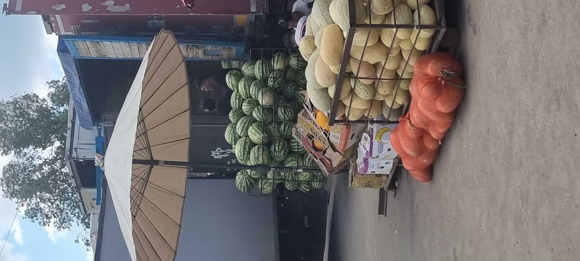 An outdoor market scene is depicted, likely in Aktobe, Kazakhstan, with a focus on a large display of watermelons and melons. A large, beige patio umbrella provides shade over a metal rack filled with numerous watermelons. Behind the watermelons, two men in dark clothing are visible, one facing away and the other in profile looking towards the watermelons. To the right of the watermelons, a metal rack is stacked high with various types of melons, some lighter green and others a yellowish hue. In the foreground, two large, bright orange mesh bags, possibly containing produce like onions or potatoes, lie on the ground. Several cardboard boxes, some with visible labels and straps, are stacked on a pallet to the left of the melons. In the background, a large truck with a blue tarp over its cargo area is parked. The ground is a rough, concrete surface. The sky above is partly cloudy.
