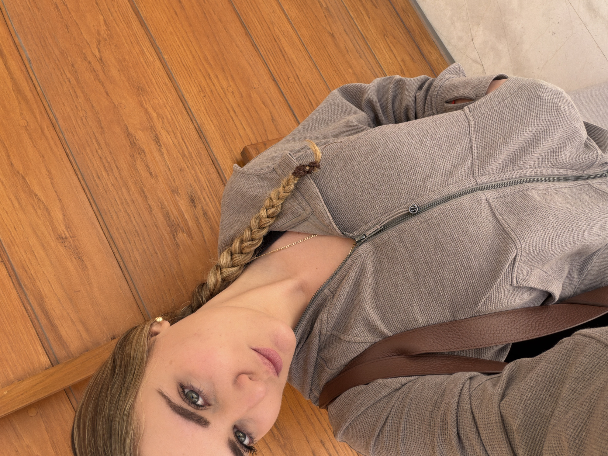 A young woman is depicted in a medium-shot portrait, reclining on a wooden floor in San Bernardino Tlaxcalancingo, Mexico. She is positioned diagonally in the frame, with her head tilted and her gaze directed towards the viewer. Her long blonde hair is styled in a side braid, adorned with a small brown hair tie, resting over her left shoulder.
She is wearing a light brown or taupe, waffle-knit zip-up jacket or hoodie, partially unzipped to reveal a delicate gold chain necklace. A small gold stud earring is visible in her left ear. A dark brown, textured leather strap, likely from a crossbody bag, runs across her chest. Her expression is neutral and engaging.
The background primarily features a warm-toned wooden floor composed of wide planks, showing natural wood grain and subtle imperfections. A small section of a lighter, possibly tiled or concrete, floor surface is visible on the far right side of the image, indicating a different flooring type or an adjacent space. The lighting is soft and appears to be natural, suggesting a daytime indoor setting. A small, indistinct symbol is visible on the zipper pull of her jacket.