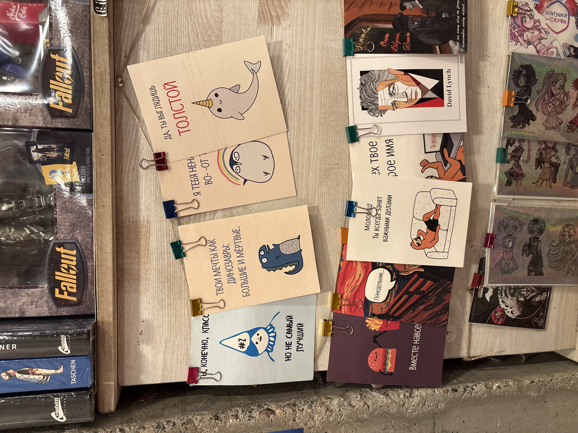 This image depicts a shelf in a store, possibly located in Saint Petersburg, Russia, based on the presence of Russian text. On the left are stacked items, including copies of "Fallout," art books from "Taschen" and "raphitti" publishing, and a "Nuka Cola" Fallout collectible. To the right, several small cards with illustrations and text in Russian are clipped to the wall, these items are affixed to the wall by clips. One card is a cartoon image of a narwhal with the word "Tolstoy" in Russian.
Another displays a drawing of a person lounging on a couch. There are also stickers. The environment is indoors and seems to be a retail space. The items are closely packed.