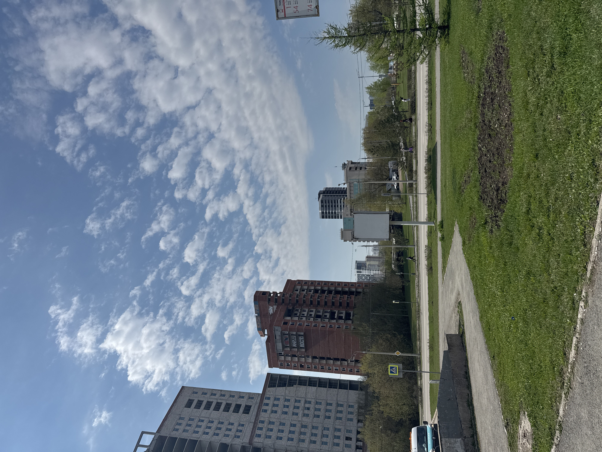 The image depicts a street scene in Perm, Russia. In the foreground is a sidewalk bordered by a strip of grass. Beyond the sidewalk, a street runs parallel, lined with trees and utility poles. In the midground, several buildings of varying heights and construction styles are visible, including a partially constructed high-rise building and a red brick building with visible graffiti. The sky, occupying the upper portion of the image, is a bright blue with scattered cumulus clouds. There are buildings on both the left and right sides of the image, indicating an urban environment. A pedestrian crossing sign is visible. There appear to be one or two distant figures walking on the sidewalk, suggesting a relatively calm urban setting.