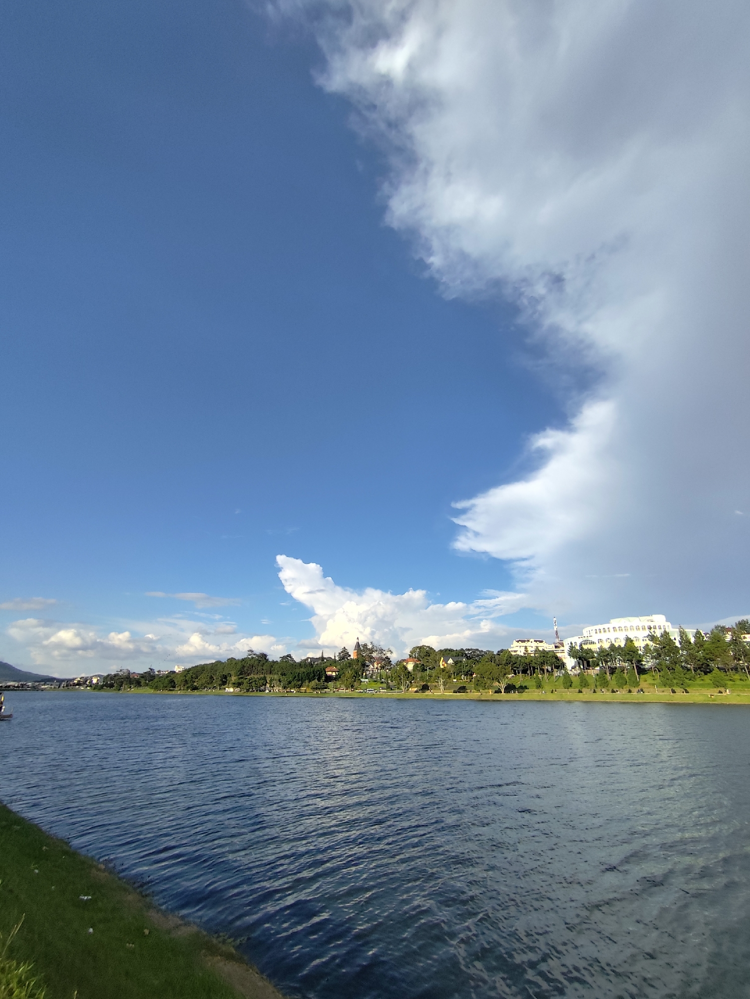 This is a wide-angle, outdoor photograph taken from the edge of a body of water, likely Xuan Huong Lake in Da Lat, Vietnam. The scene depicts a clear blue sky with scattered white clouds on the left, transitioning to darker, stormier clouds on the right.
In the foreground, the rippling surface of the water occupies the majority of the lower frame. The water is a deep blue and reflects the sky. To the immediate left of the water is a strip of green grass, with some white specks, possibly flowers or debris, scattered on it.
The middle ground features a lush green shoreline with a line of trees. Beyond the trees, a cluster of buildings and structures are visible on a gentle incline. These include several multi-story buildings, some with white facades and architectural details like arched windows, and at least one structure with a pointed roof, possibly a church spire. A tall, slender radio tower is also present among the buildings.
The background is dominated by the sky. The contrast between the bright blue sky with wispy clouds on the left and the heavy, gray clouds on the right suggests an approaching weather front or a shift in atmospheric conditions. The overall impression is a natural landscape blended with urban development, bathed in natural light under a dynamic sky. No people are clearly discernible as distinct subjects in the image, though there are hints of human activity on the distant shoreline.
