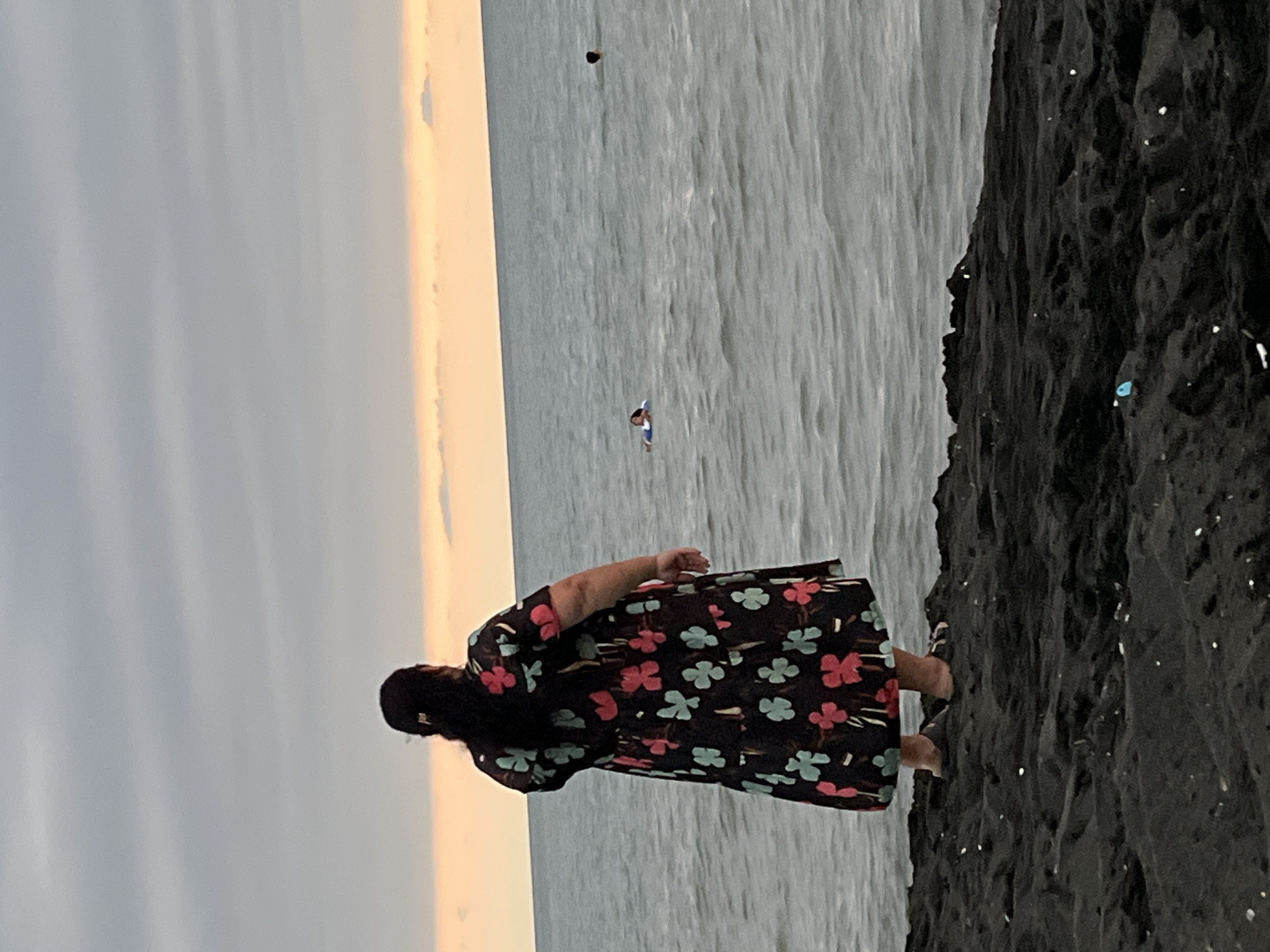An overcast sky looms above a choppy sea and a dark, sandy shore. A woman with dark hair, wearing a patterned dress and sandals, stands facing the water. In the sea, a person floats on a blue raft, and further out, another person's head is visible. The water is grey and wavy, reflecting the dull sky. The shore is covered in dark sand, with some small white specks scattered across it. The scene appears to be at the Black Sea coast of Ureki, Georgia, a town known for its magnetic black sand beaches.