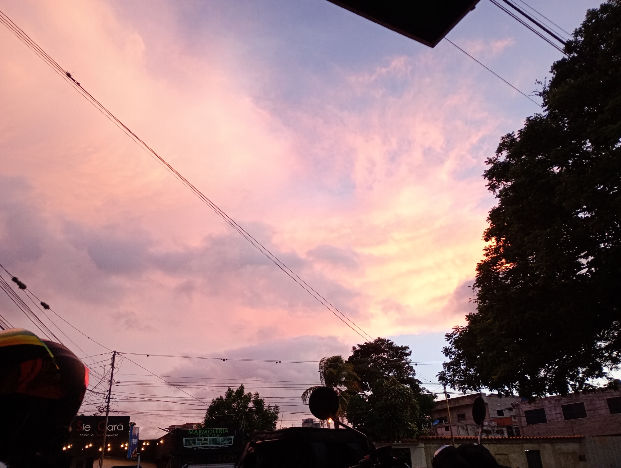 The image shows a street scene in Barquisimeto, Venezuela, at dusk. The sky is filled with pink and purple clouds. Power lines crisscross the sky. In the foreground, there is a motorcycle, possibly being ridden. In the background, there are trees, buildings, and commercial establishments, including signs for "Sie Cara" and "Marmoleria Tecnito". The overall impression is of a city street at sunset.
