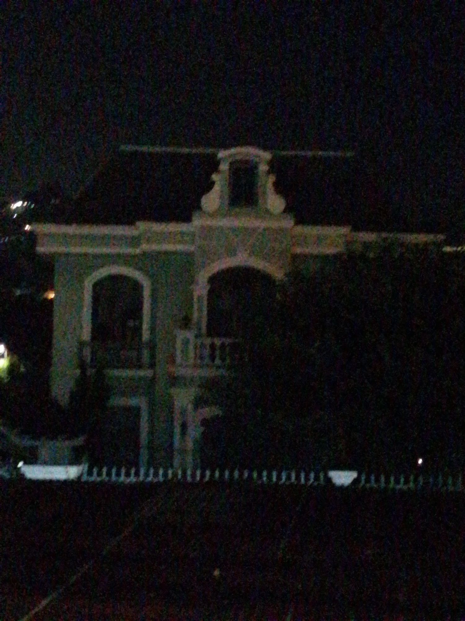 The image shows a dimly lit, slightly out-of-focus view of a two-story building, possibly a house or a small hotel, at night. The building has a distinctive architectural style, featuring arches above doorways and windows, and decorative elements along the roofline. The main facade appears to be a pale green color. There are dark trees or foliage obscuring parts of the building and the background. In the foreground, there is a fence or railing, which is also dimly visible. The overall impression is a residential or commercial property in an urban or semi-urban setting, likely in Da Lat, Vietnam, given the architectural style often found in the city, though this cannot be definitively confirmed from the image alone. There are no people visible in the image.