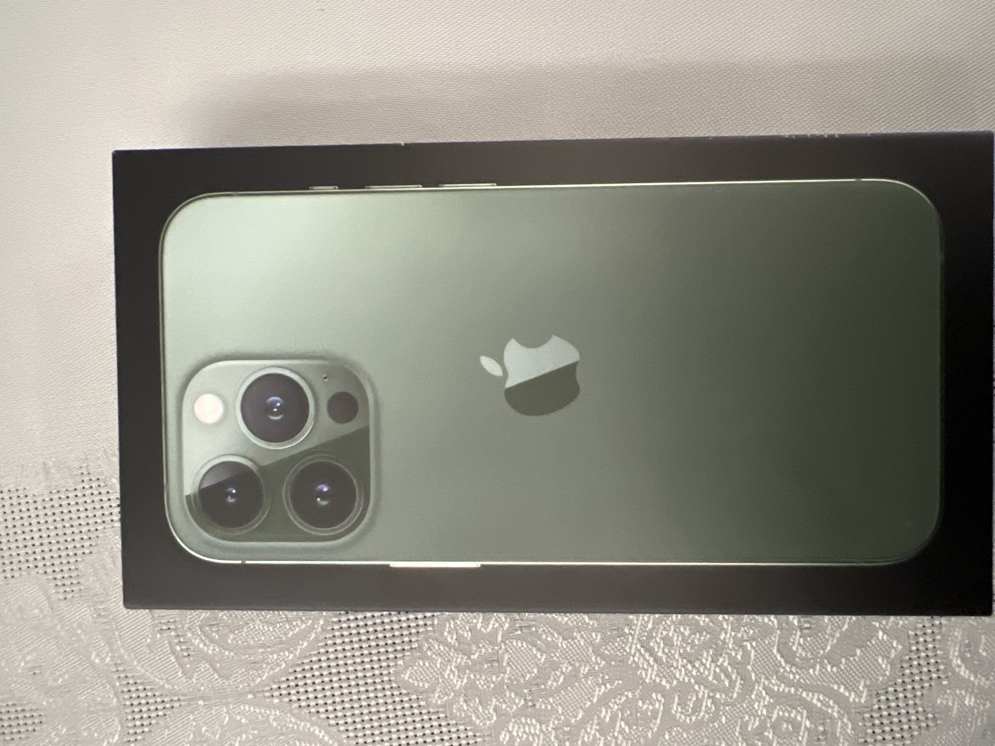 The image shows a closed retail box of an Apple iPhone. The iPhone depicted on the box is green in color with three camera lenses and an Apple logo. The box is black. The surface beneath the box appears to be a textured, possibly patterned, white or light-colored surface. The photo does not contain sufficient information to determine the location within Burgas, Bulgaria, or to provide further details about the environment, people, actions, or interactions.