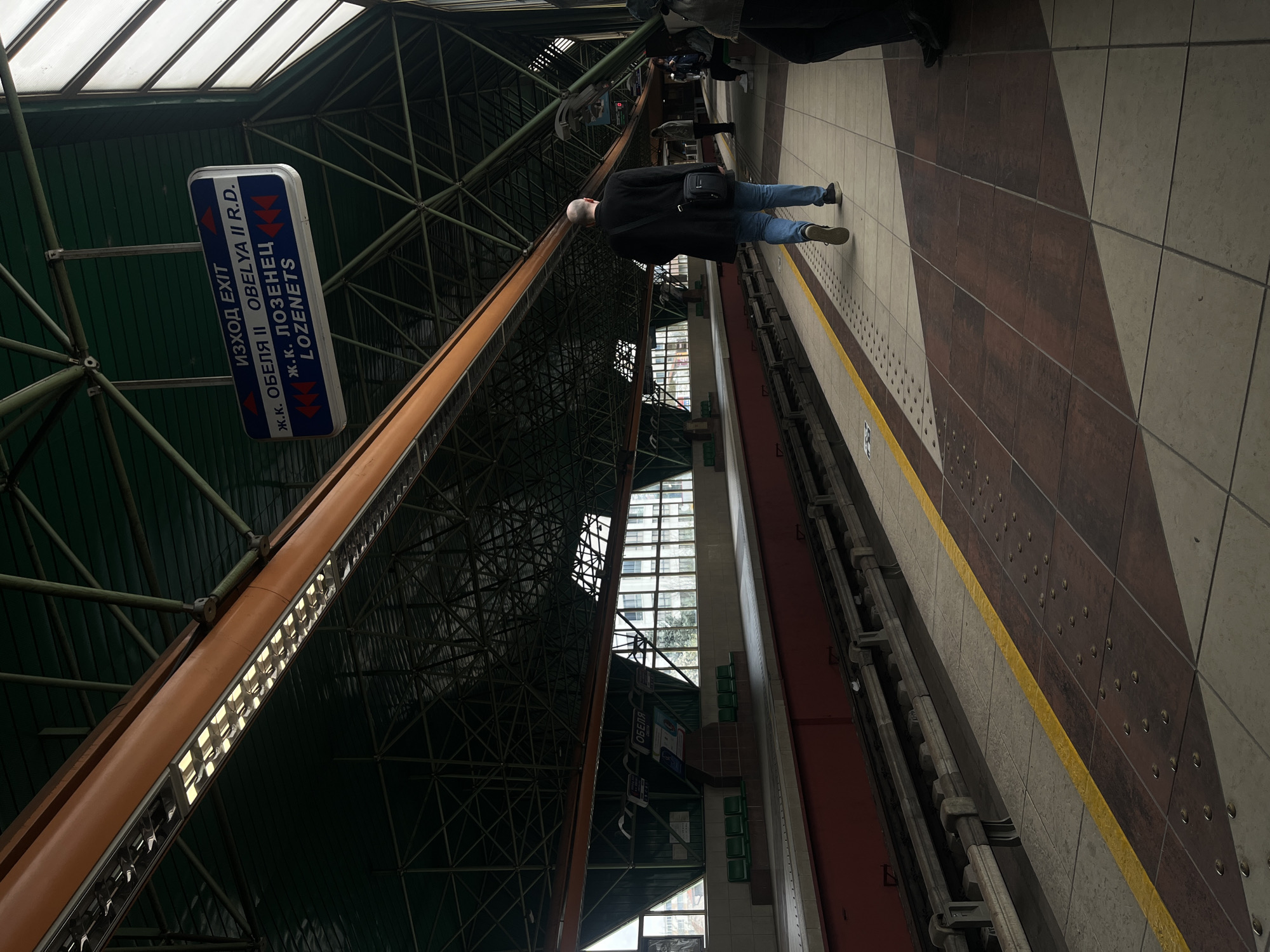 The image shows the interior of a metro station in Sofia, Bulgaria. The station is characterized by a high, angled ceiling with exposed steel framework and a green-painted upper surface. A large, vertical sign indicates "EXIT" in both Bulgarian and English, along with directions to "OBELYA II R.D." and "LOZENETS". A middle-aged man is standing on the station platform, wearing a black coat, blue jeans, and dark shoes. He carries a black shoulder bag. A yellow safety line is painted along the platform edge. The platform has a pattern of brown and beige tiles. The overall lighting in the station is bright due to the skylights and overhead lighting fixtures. Other individuals are faintly visible in the background.