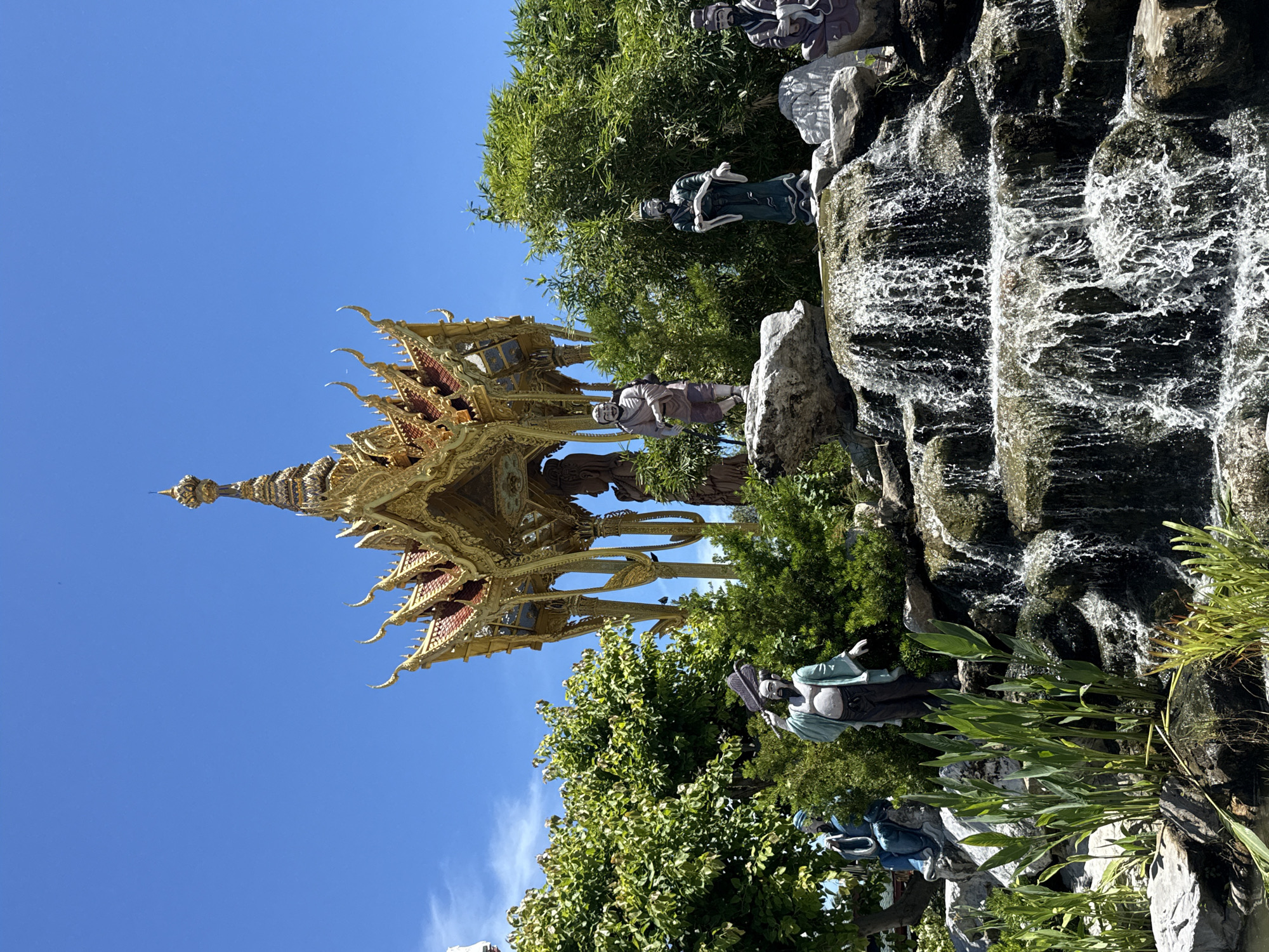 The image depicts a landscape in Bang Pu Subdistrict Municipality, Thailand, featuring a golden pavilion-like structure juxtaposed against a rock waterfall and verdant vegetation. The pavilion is ornately decorated with intricate gold and red detailing. Numerous statues of figures in traditional Chinese dress are placed among the greenery and rocks. The waterfall cascades down dark-colored rocks, its water appearing foamy and white. The sky is a clear blue.