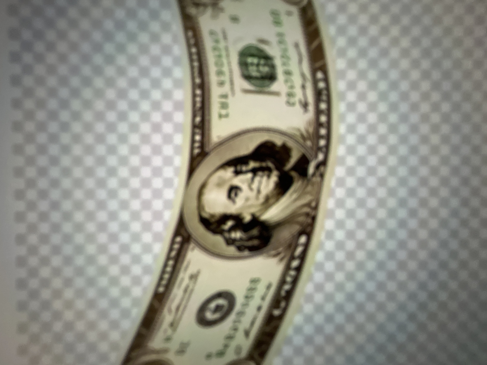The image displays a close-up, distorted view of a U.S. dollar bill. The bill is curved and appears to be on a transparent, checkered background. The portrait of Benjamin Franklin is visible on the left side of the bill. The denomination "TWENTY" is partially visible at the top left and top right. The number "20" is also present on the bill. Due to the blurriness and distortion, specific details like serial numbers or fine print are illegible. The image quality is poor, making precise identification challenging. There are no people, actions, interactions, or elements related to the city of Gelendzhik, Russia, present in this image.