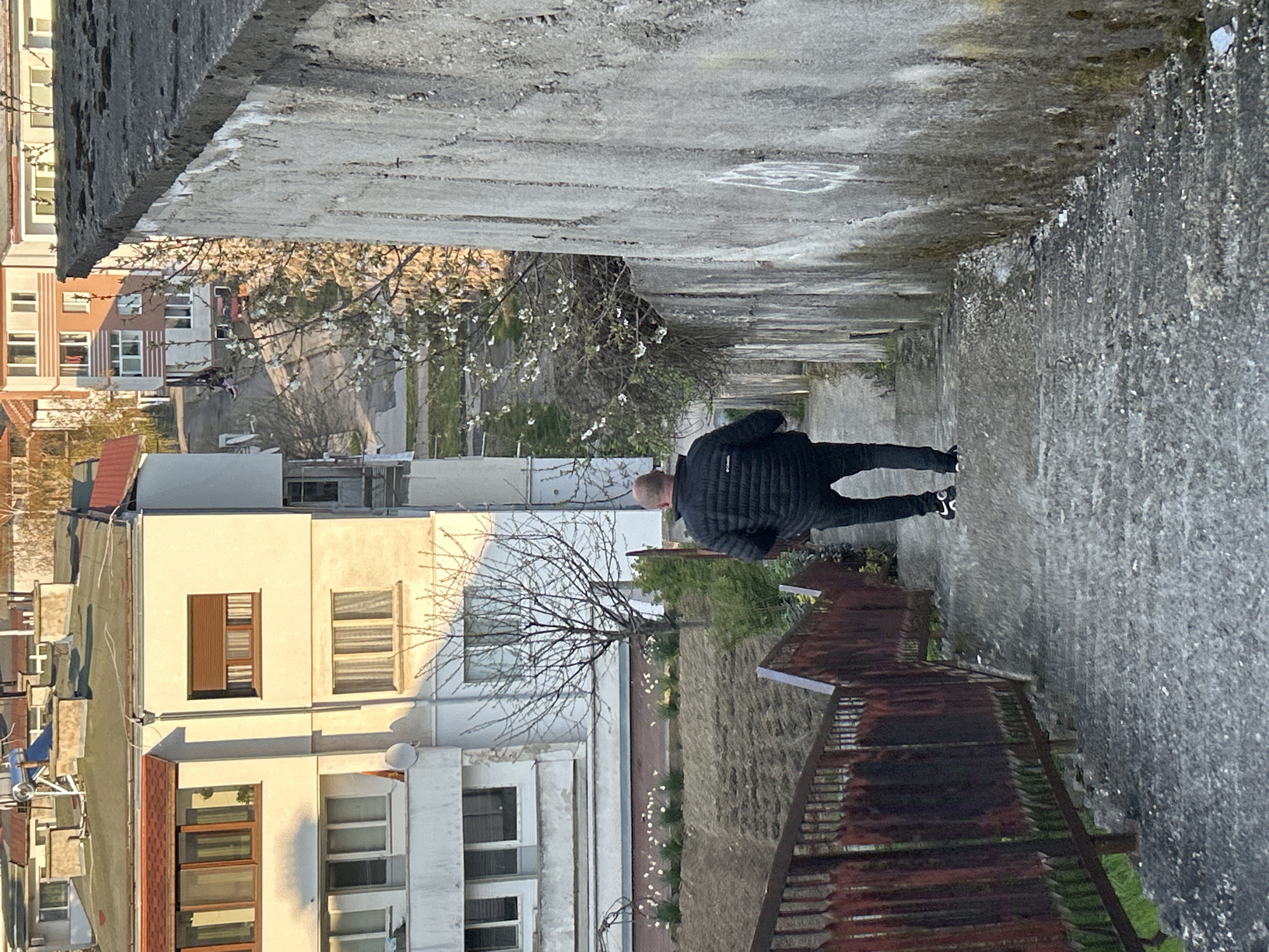 The image shows an individual walking on a sidewalk in Zlatograd, Bulgaria. The scene is captured from an elevated perspective, looking down on a narrow passageway between two buildings. The individual is wearing a dark jacket and dark pants, walking away from the viewer. A tree is located to the side of the sidewalk. The building on the left side of the image is a multi-story building with white walls and various window designs. A rusty metal fence is located on the right side of the sidewalk. Above the sidewalk is a concrete structure, possibly the underside of a bridge or overpass. The sidewalk appears to be concrete as well.