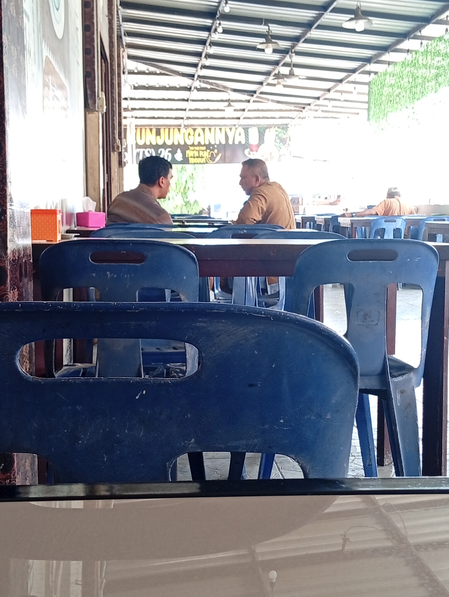 The image captures an indoor/outdoor seating area, likely a restaurant or cafe, with a covered roof supported by a metal frame. Two men are seated at a table, facing each other and engaged in conversation. The man on the left is wearing a brown shirt and looking to his right. The man on the right is also wearing a brown shirt and looking towards the man on the left. In the foreground, several blue plastic chairs are visible, some occupied and others empty. Behind the seated men, a sign with Indonesian text, including "UNJUNGANNYA," is partially visible. The environment appears to be a public space with a casual atmosphere. The scene is likely located in Lhokseumawe, Indonesia, as indicated by the presence of Indonesian text on the sign and the typical architectural style of such establishments in the region. The lighting suggests it is daytime, with natural light filtering through the roof and the open sides of the structure.