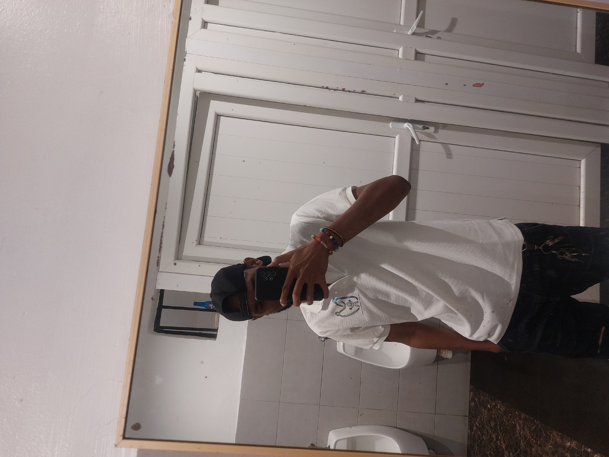 A man is taking a selfie in a mirror in what appears to be a public restroom. The man is wearing a white collared shirt, dark pants, and a black baseball cap. He is holding a black smartphone in front of his face. The restroom has white tiled walls and a white tiled floor. There are two white urinals visible in the mirror. Behind the man is a white door with horizontal panels. There is also a black metal shelf visible in the mirror. Without additional information, it is not possible to determine the location of the restroom in Dar es-Salaam, Tanzania.