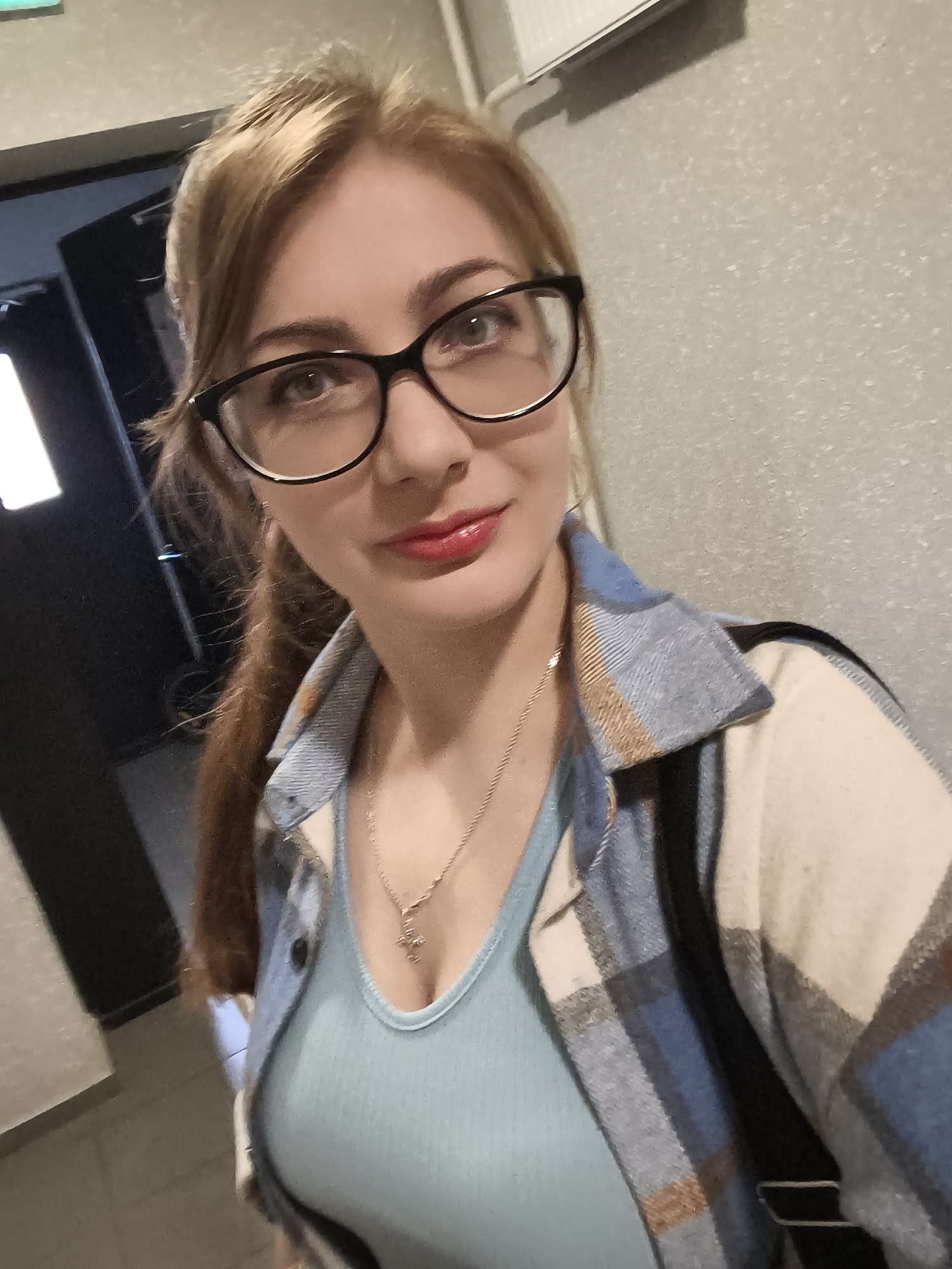 A woman with blonde hair and glasses is taking a selfie in an indoor setting. She is wearing a light blue ribbed tank top and a plaid shirt over it. The background appears to be a hallway with a textured wall and a dark doorway. She is looking at the camera with red lipstick.