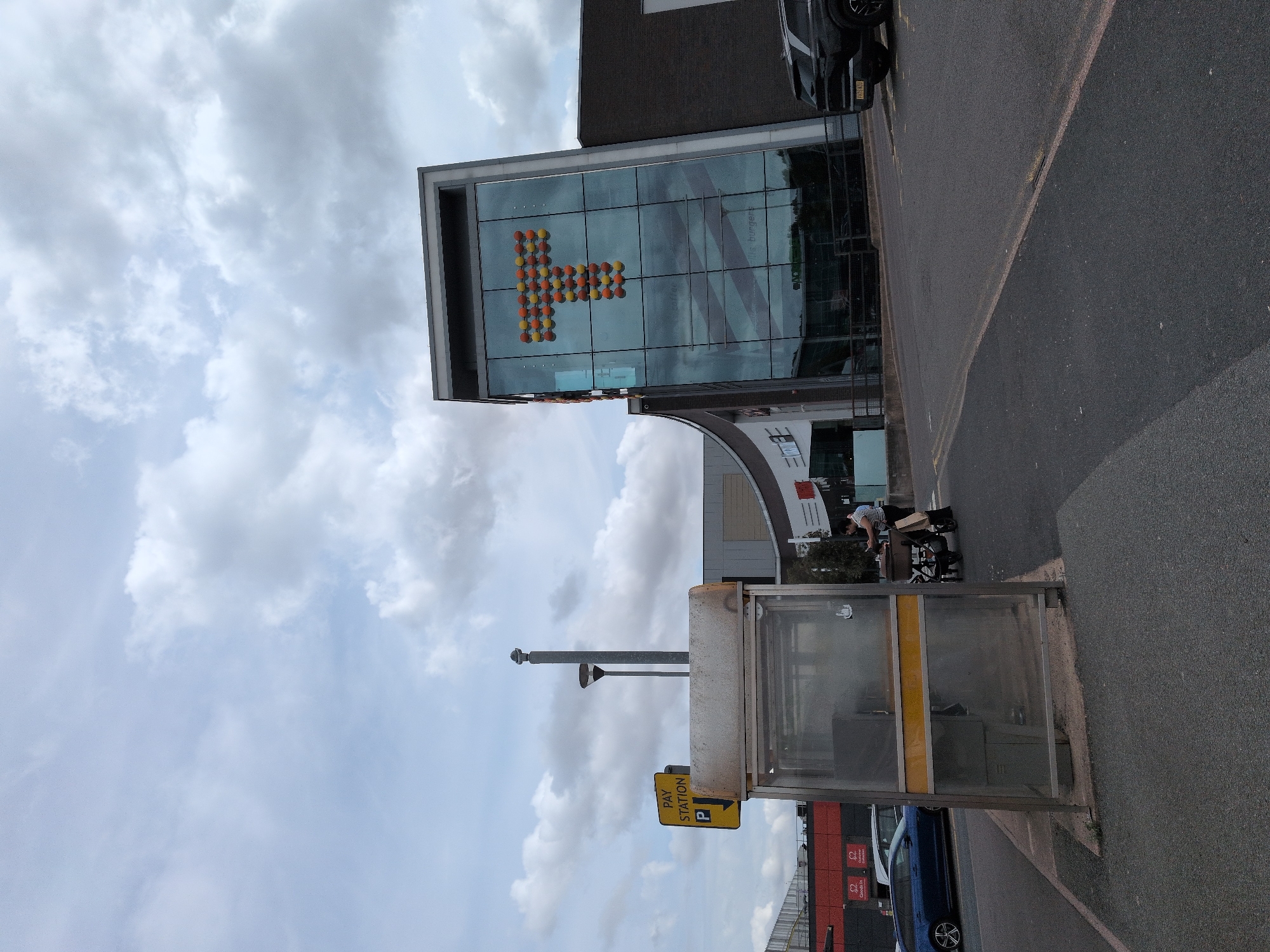 The image shows a scene outside a shopping center in Telford, United Kingdom. In the foreground, there is a pay station booth, made of metal and glass, with a yellow sign above it indicating "PAY STATION" and a parking symbol. To the right of the pay station, a woman is pushing a stroller. Behind the pay station, a dark blue car is parked on the left side of the road, and a black SUV is parked on the right side. In the background, the shopping center building is visible, featuring a large glass facade adorned with a colorful "T" made of spheres. The sky is cloudy.
