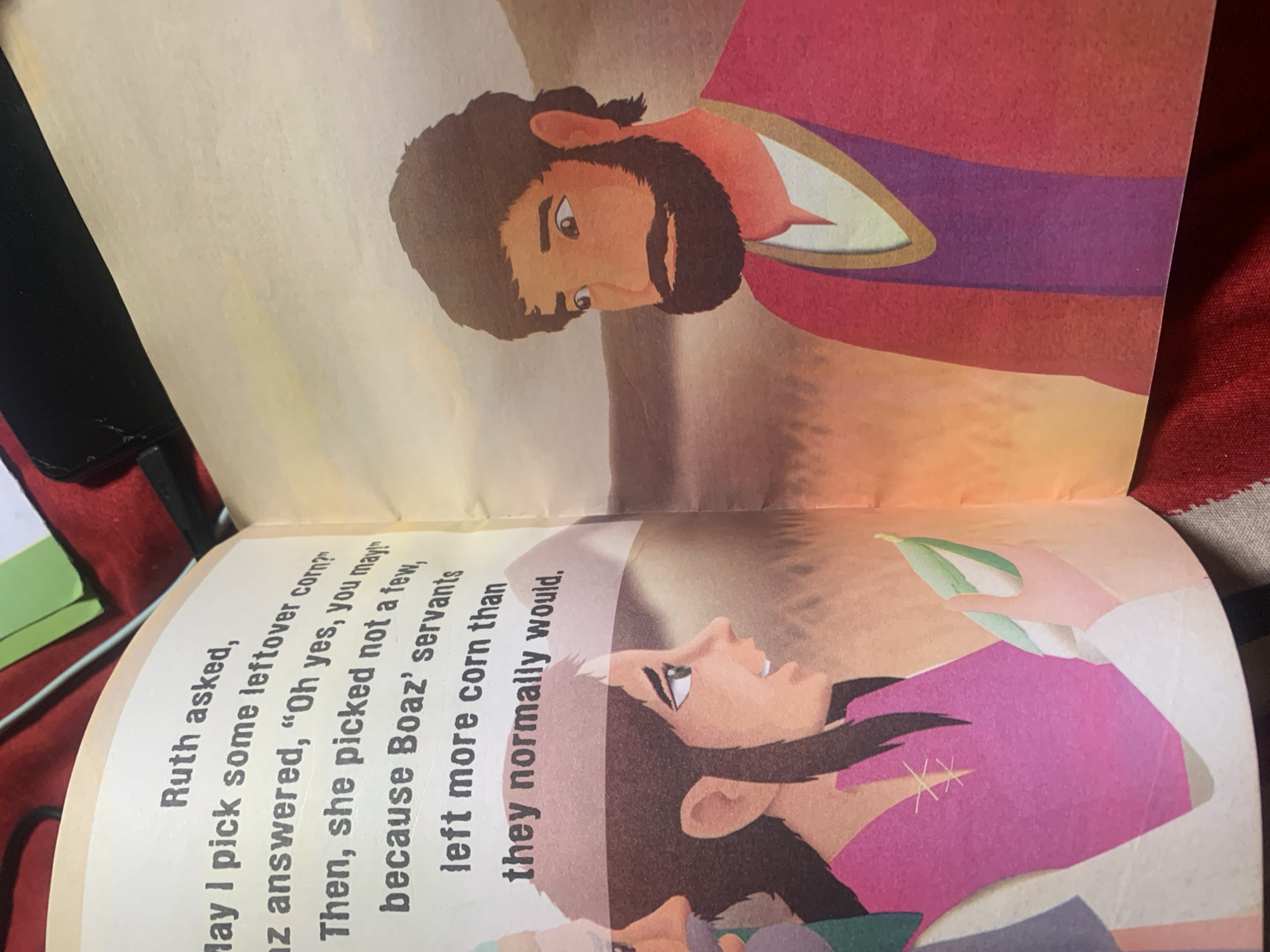 The image shows an open book with an illustration and text. The illustration depicts two people, a woman and a man, in a pastoral setting. The woman, on the left, is wearing a pink tunic and has dark hair. She is holding a few green pods in her hand. To her right is a man with a beard and dark hair, wearing a red robe. The text on the left page reads: "Ruth asked, May I pick some leftover corn? az answered, "Oh yes, you may!" Then, she picked not a few, because Boaz' servants left more corn than they normally would." There is no information in the image to suggest it is located in Jauro Yinu, Nigeria.