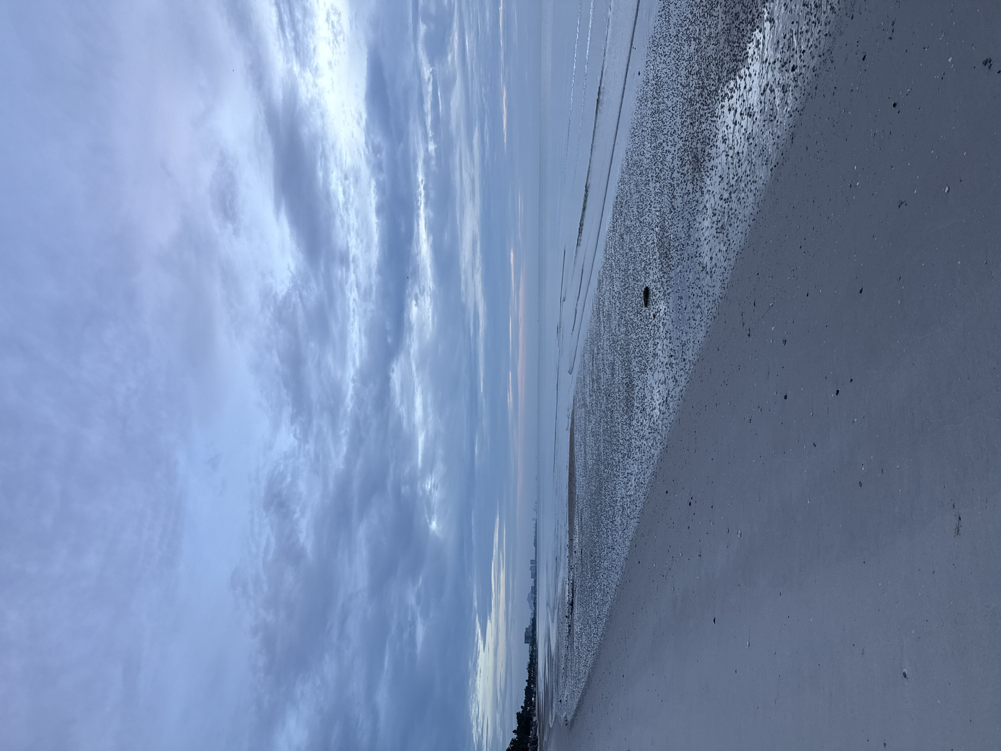 The image depicts an overcast morning on a beach in Hua Hin, Thailand. The sky is filled with a dense layer of grey and pale blue clouds, with some breaks allowing for subtle hints of pink and orange light near the horizon, suggesting the early stages of sunrise or sunset. The sea is calm, with gentle waves lapping at the shore. The beach itself is composed of wet sand and a scattering of small dark pebbles or shells, particularly in the intertidal zone. In the distance, a hazy cityscape is visible along the coastline, with several buildings, including hotels and residential structures, emerging from the mist. The overall atmosphere is tranquil and subdued.
