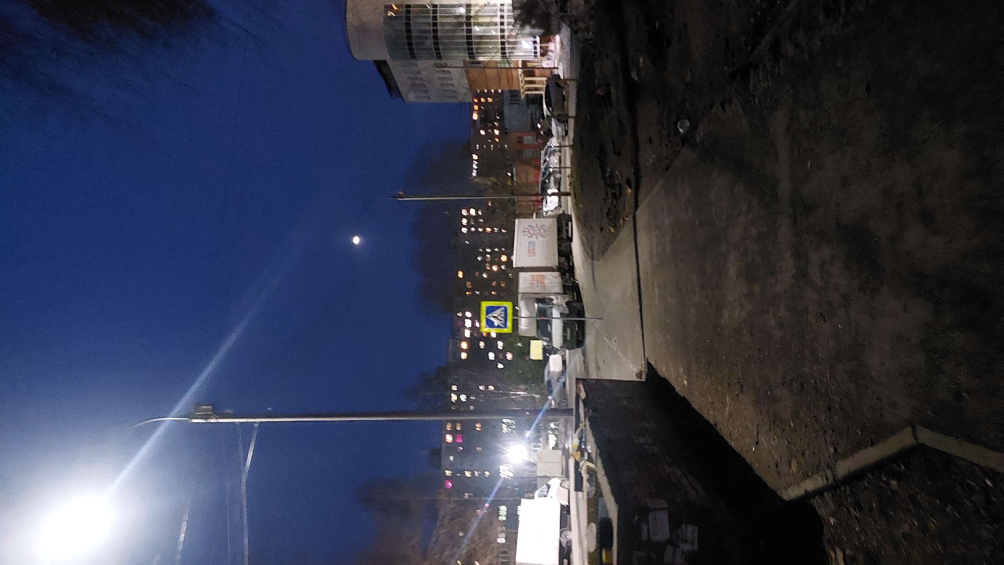 The image depicts a street scene in Novosibirsk, Russia, at night. The sky is dark blue with a visible moon. A street lamp illuminates the lower part of the frame. Several buildings with lit windows are visible in the background, indicating an urban setting. A yellow and blue pedestrian crossing sign is present along the street. Several vehicles, possibly trucks, are parked along the side of the street. The foreground features a sidewalk with a dark, uneven surface.