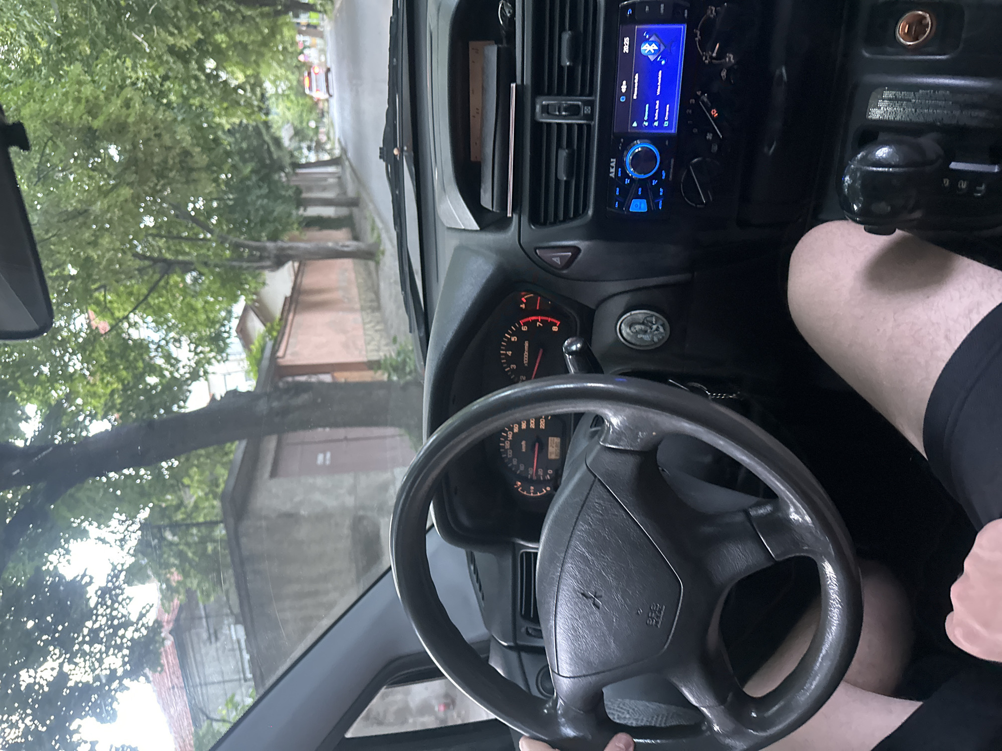 The image is a first-person view from inside a car, looking forward. The steering wheel, dashboard with gauges and an infotainment screen, and gear shifter are visible. The driver's legs are also in the frame. Outside the car, a tree-lined street with houses on either side is visible. A white car is seen in the distance. The lighting suggests it is daytime. The image appears to be taken while the car is in motion.