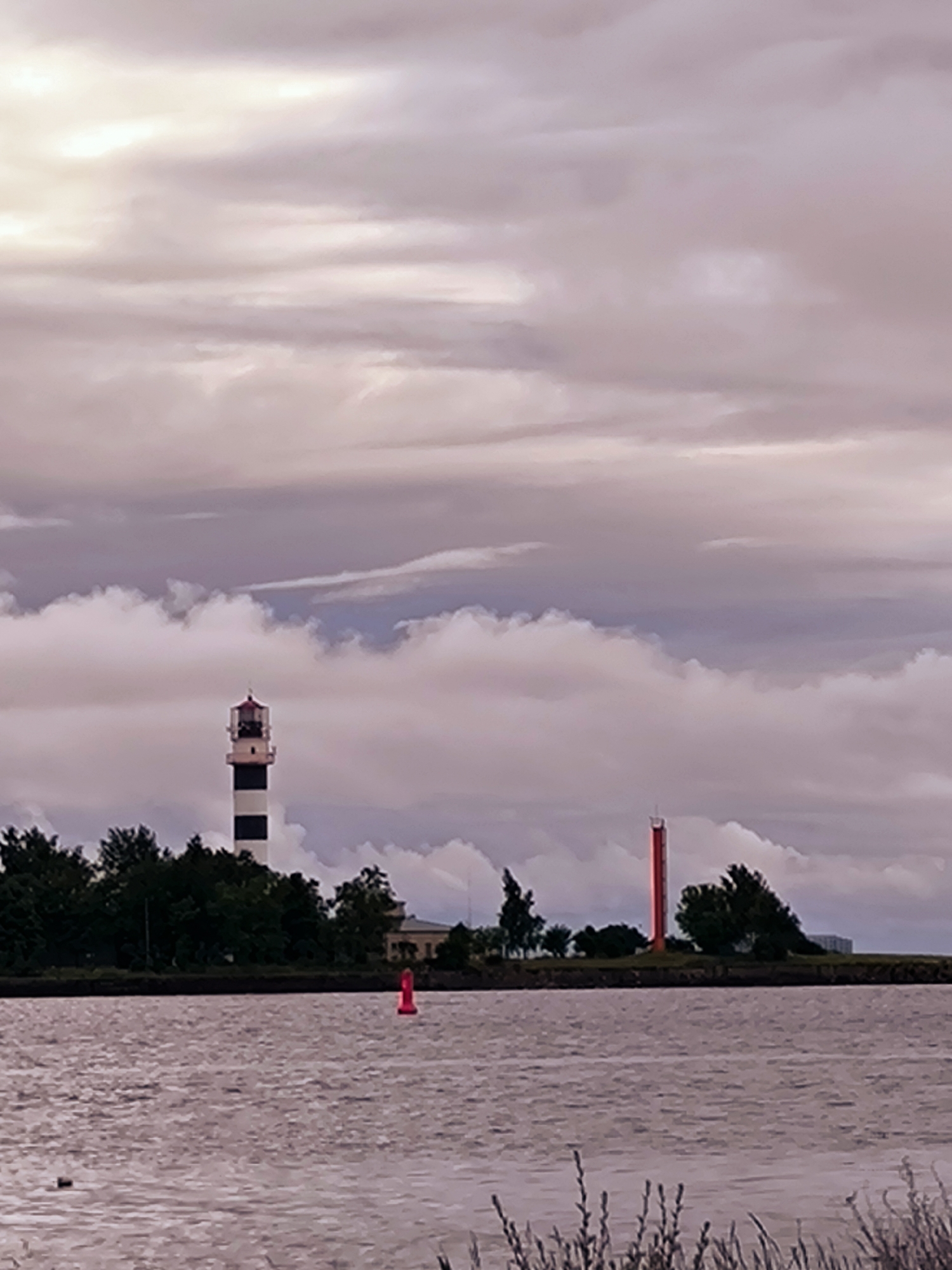 This image depicts a wide body of water with a shoreline in the background, under a cloudy sky. On the left side of the shoreline, a striped lighthouse, black and white, stands tall. To the right of the lighthouse, a red, tall, slender tower is visible. Trees line the shoreline, and in the water, a red buoy is present. The scene is likely a port or coastal area, possibly near Riga, Latvia, given the presence of maritime structures like lighthouses and buoys. The overall mood is somber, with the overcast sky casting a muted light on the scene.