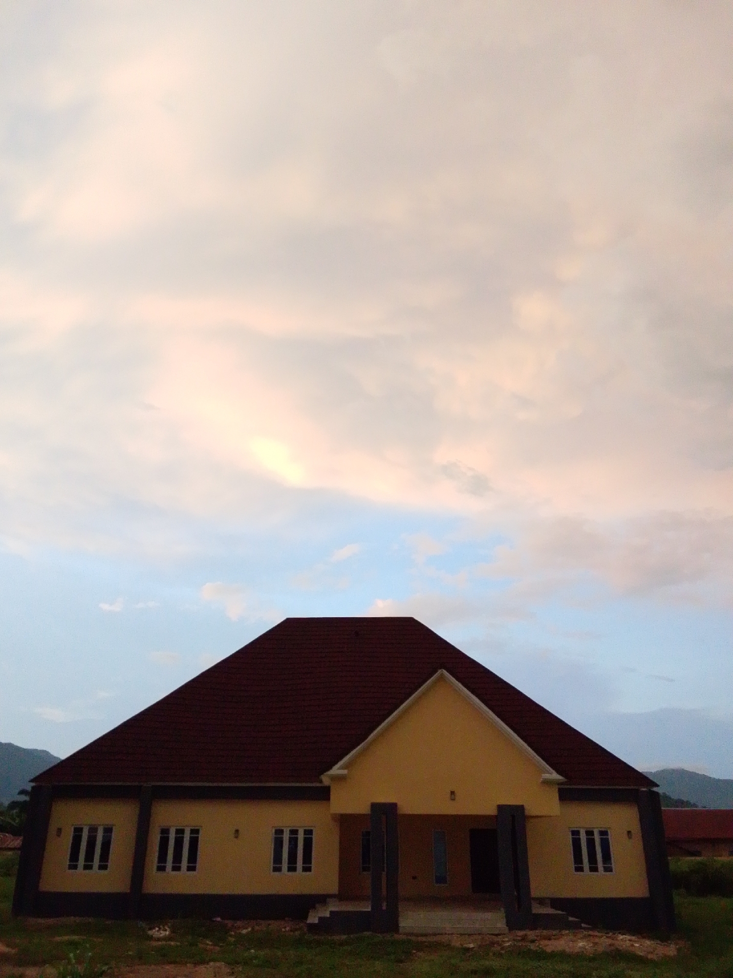 This is a photograph of a one-story building with a dark red, multi-pitched roof, located in Serti, Nigeria. The building's facade is painted in a light yellow color, with darker brown trim around the base and windows. There are multiple rectangular windows with white frames and visible panes. The building appears to be situated in an open area with some dry grass and soil in the foreground. In the background, there are hints of hilly terrain and possibly other structures. The sky above is filled with clouds, exhibiting a mix of pale pink and light blue hues, suggesting it may be either sunrise or sunset.