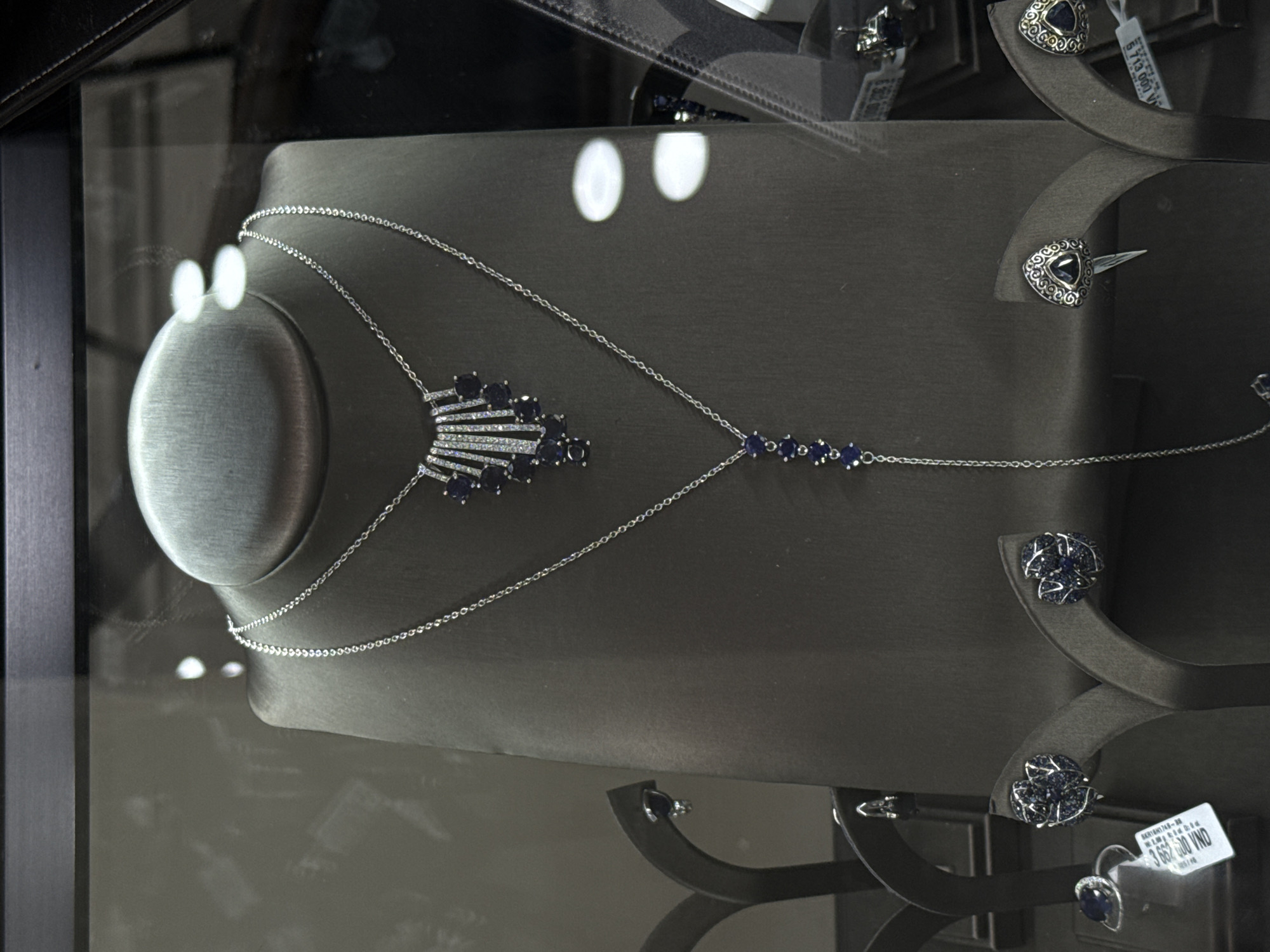 A jewelry display in Nha Trang Ward, Vietnam, features a gray mannequin displaying a silver necklace with dark blue gemstone accents. Several pairs of silver earrings with dark blue gemstones are positioned on small gray stands surrounding the mannequin. Price tags are visible on the earrings. The display is behind glass. The lighting is bright, with reflected spots on the glass.