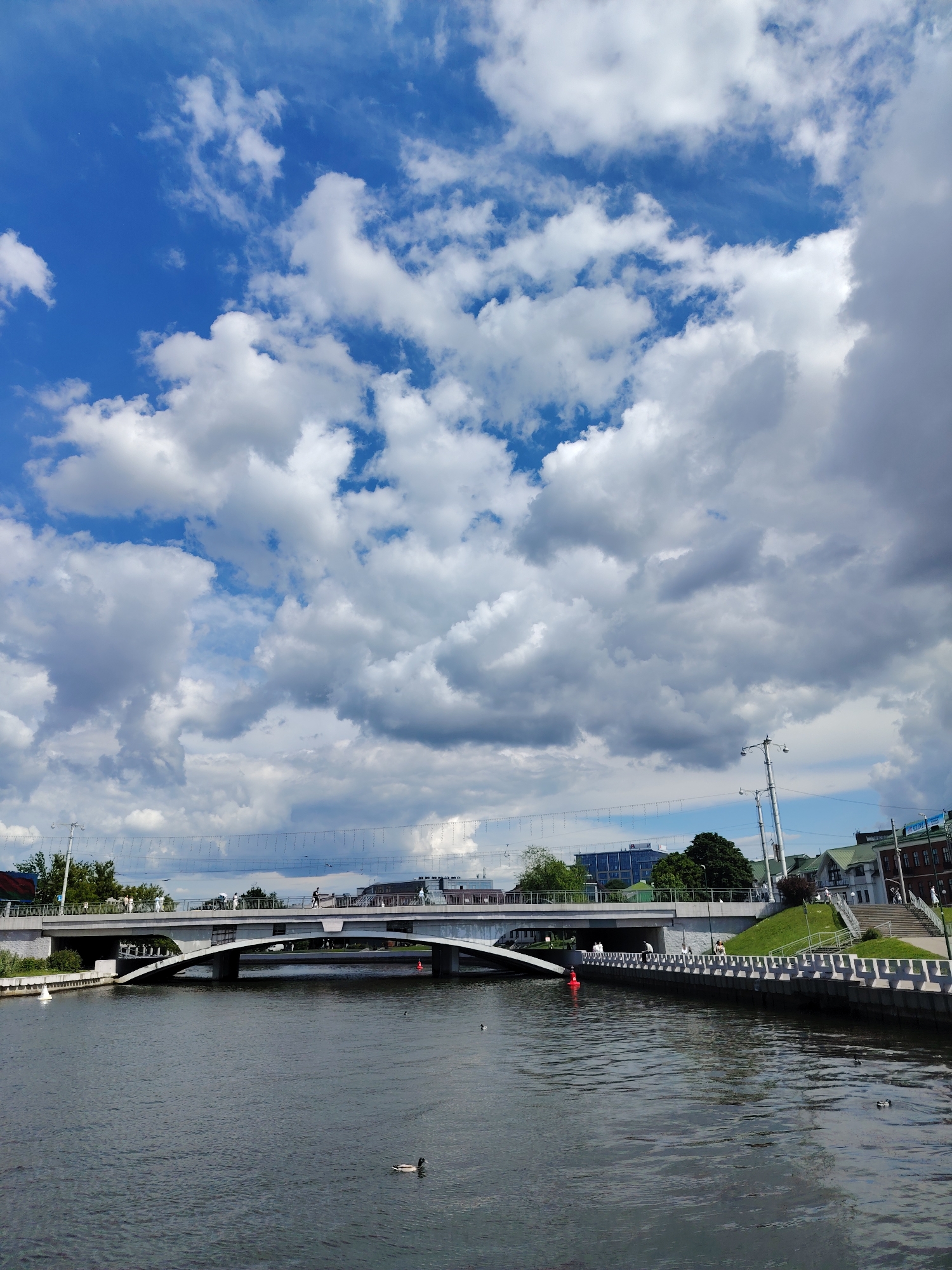 This image captures a vibrant daytime scene along a river or canal in Minsk, Belarus, under a striking sky. The upper portion of the frame is dominated by a clear blue sky interspersed with large, fluffy white and grey cumulus clouds, indicating a dynamic weather pattern with both sun and cloud cover.
The central focus is a modern, light-colored concrete bridge with a gracefully arched underside, spanning the calm, dark waters of the river. Several people are visible walking across the bridge, and others can be seen along the riverbanks, suggesting everyday urban activity. On the left bank, there are some contemporary buildings and greenery. The right bank features a robust stone or concrete embankment, alongside which are lush green grassy slopes and a series of steps leading up to older-style buildings with distinctive green roofs.
A bright red buoy stands out in the water near the right bank, and a white buoy is situated closer to the left. A few ducks are seen swimming in the river, adding to the tranquil atmosphere. Decorative string lights or wires are suspended above the bridge, stretching across the sky, and various streetlights and utility poles are also visible. The scene portrays a bustling yet peaceful urban environment, showcasing a blend of architecture, nature, and daily life. No visible text is discernible in the image.