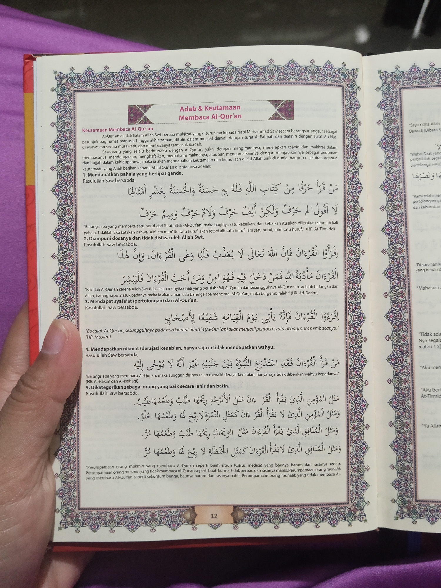 The image shows an open book, likely a religious text, with decorative borders. The text is in Arabic and Indonesian. On the left page, there is a section titled "Adab & Keutamaan Membaca Al-Qur'an" (Etiquette & Virtues of Reading the Qur'an). The text lists five points related to the benefits and virtues of reading the Qur'an, with some verses in Arabic script and explanations in Indonesian. On the right page, there are quotes in Arabic and Indonesian, possibly related to the same religious text, and decorative borders. The book is open on a surface, and part of a hand is visible in the lower left corner, holding the book open. The background is out of focus but suggests an indoor setting, possibly a room with a purple cloth draped. The lighting appears to be natural or artificial, casting soft shadows. There are no discernible landmarks or features to indicate a specific location in the city of Lestari Indah, Indonesia. The image focuses on the book and its content.