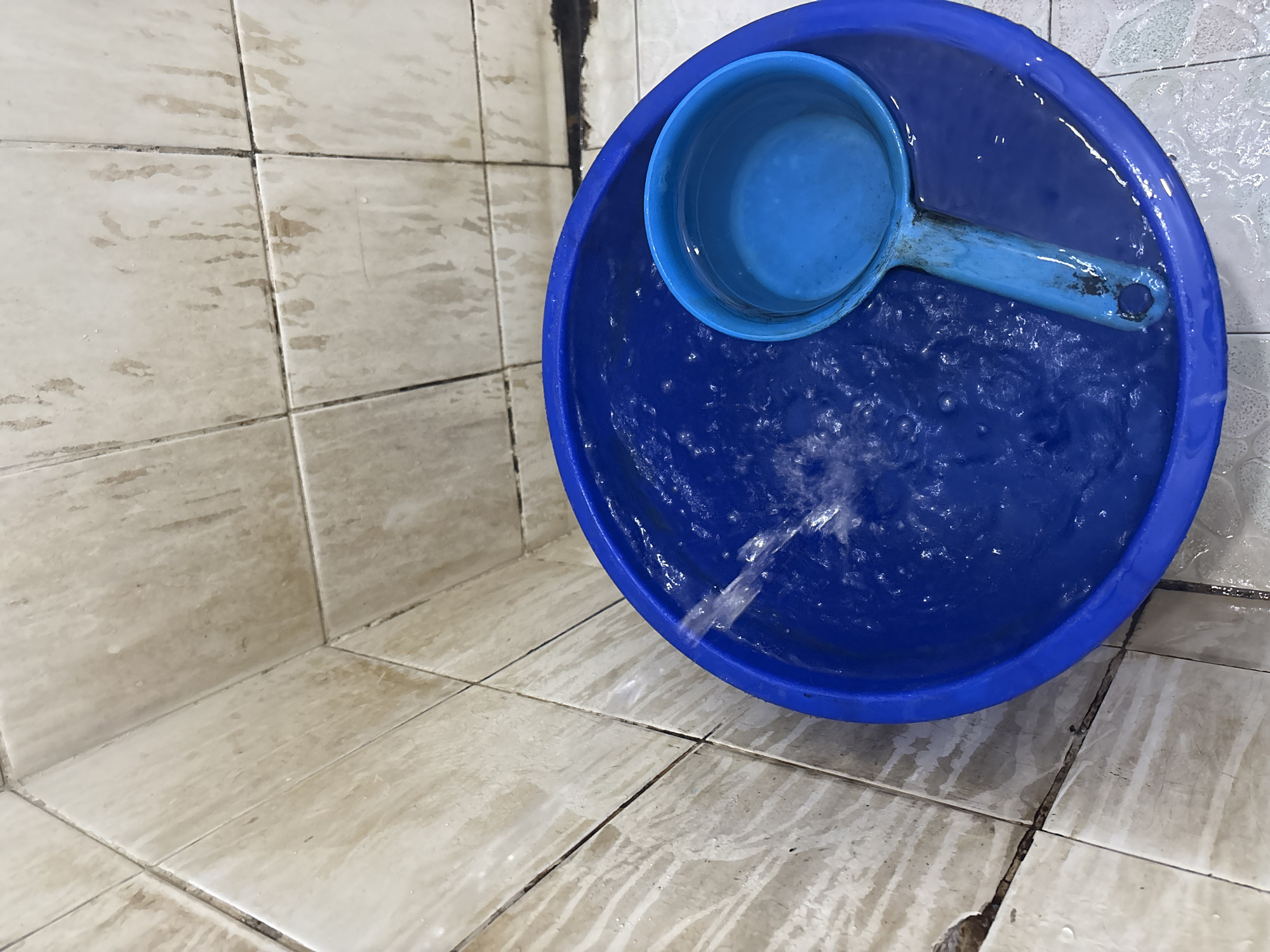 In Palimanan, Indonesia, a bright blue plastic bucket is being filled with water, which streams from an unseen source. Inside the bucket, a smaller blue plastic ladle rests, partially submerged in the swirling water. The bucket is positioned on a tiled floor, adjacent to a tiled wall. The tiles appear to be a light beige color and show signs of wear and possibly mildew in the grout lines. The overall scene suggests a practical, possibly domestic, setting where water is being collected.