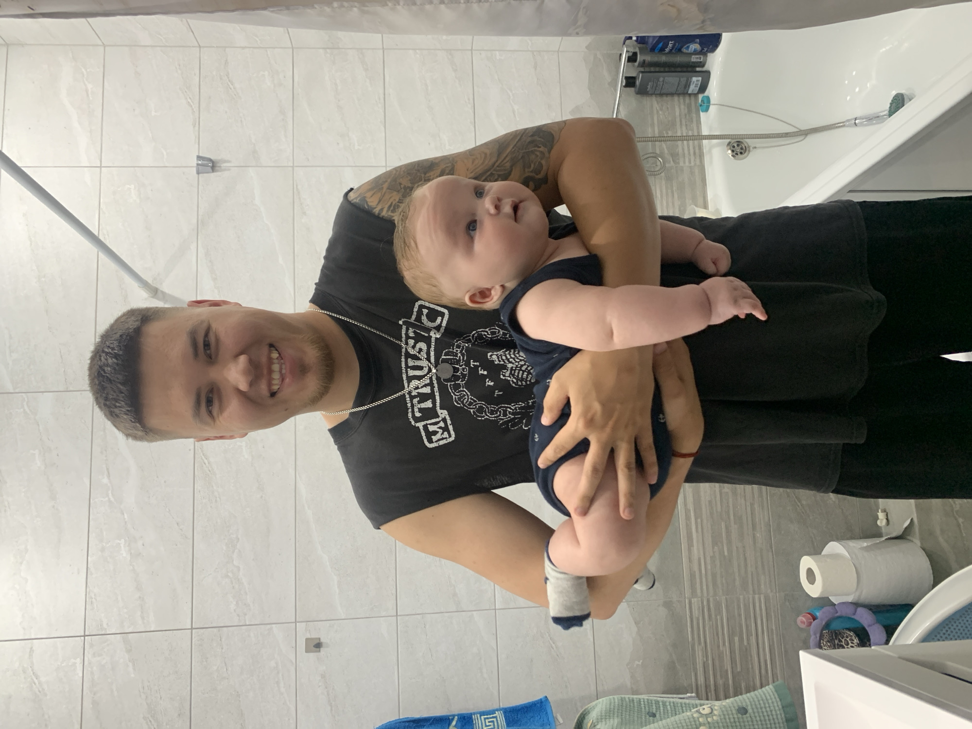 In a bathroom with light-colored tiled walls, a young adult male with short, dark hair and a clean-shaven face smiles at the camera while holding a baby. He is wearing a black, sleeveless t-shirt with a graphic design and a silver chain necklace. The baby, who is facing to the right and looking away from the camera, is wearing a dark blue onesie and white socks. The baby’s arms and legs are visible and appear chubby. Behind them, a bathtub with a shower curtain is partially visible, along with toiletries on a shelf above the tub. To the left, a sink and a roll of toilet paper are present. The overall scene suggests a casual, domestic moment. No specific landmarks of Stavropol, Russia are discernible in the image.