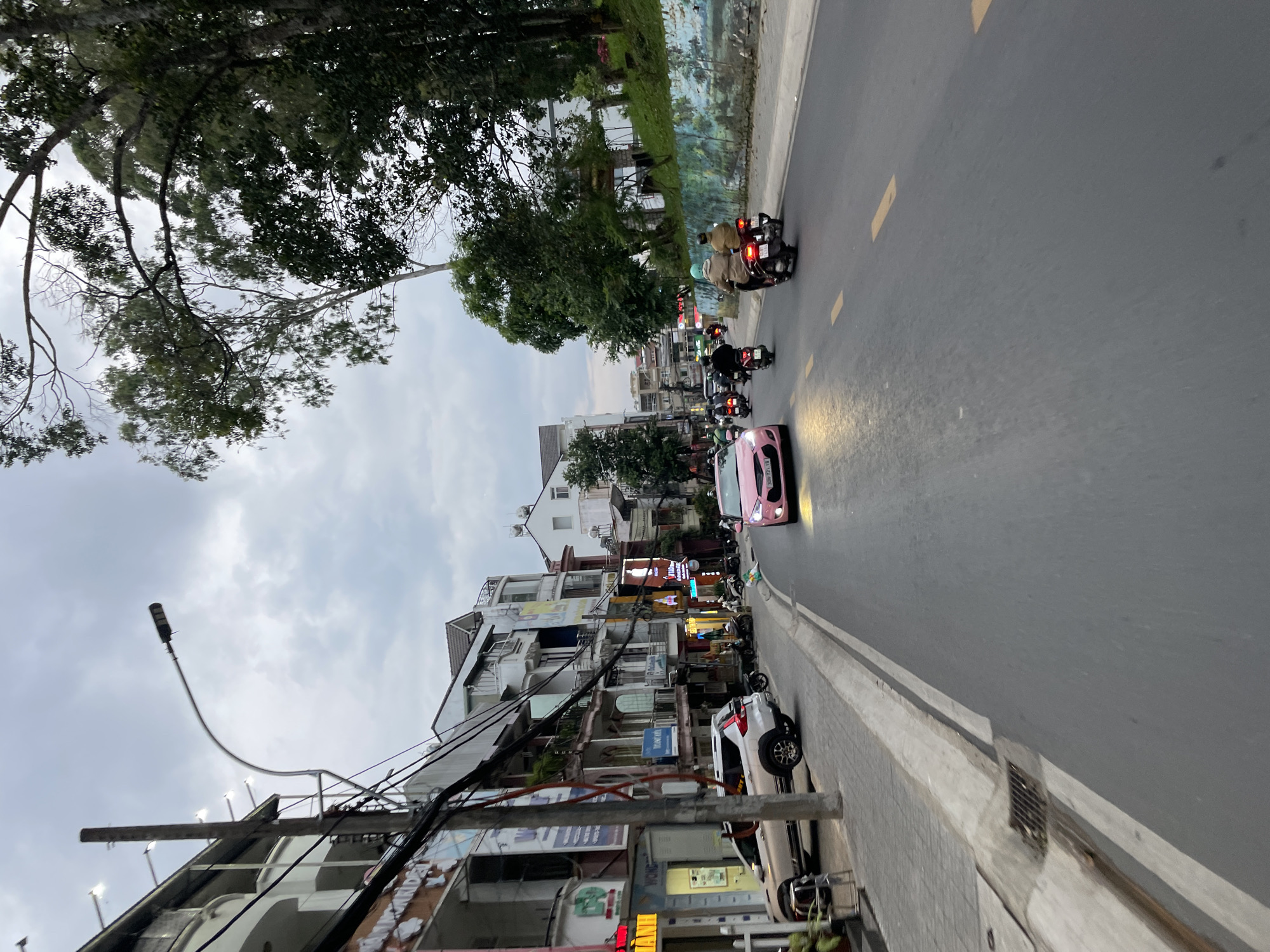 The image depicts a street scene in Da Lat, Vietnam. A pink car is in the foreground, driving on the right side of the road. Several motorcycles are also visible, some carrying passengers. Buildings line the left side of the street, with shops and apartments. Trees and vegetation are present on the right side of the road, with a mural painted on a retaining wall. The sky is overcast. The overall impression is a typical street in a Southeast Asian city.