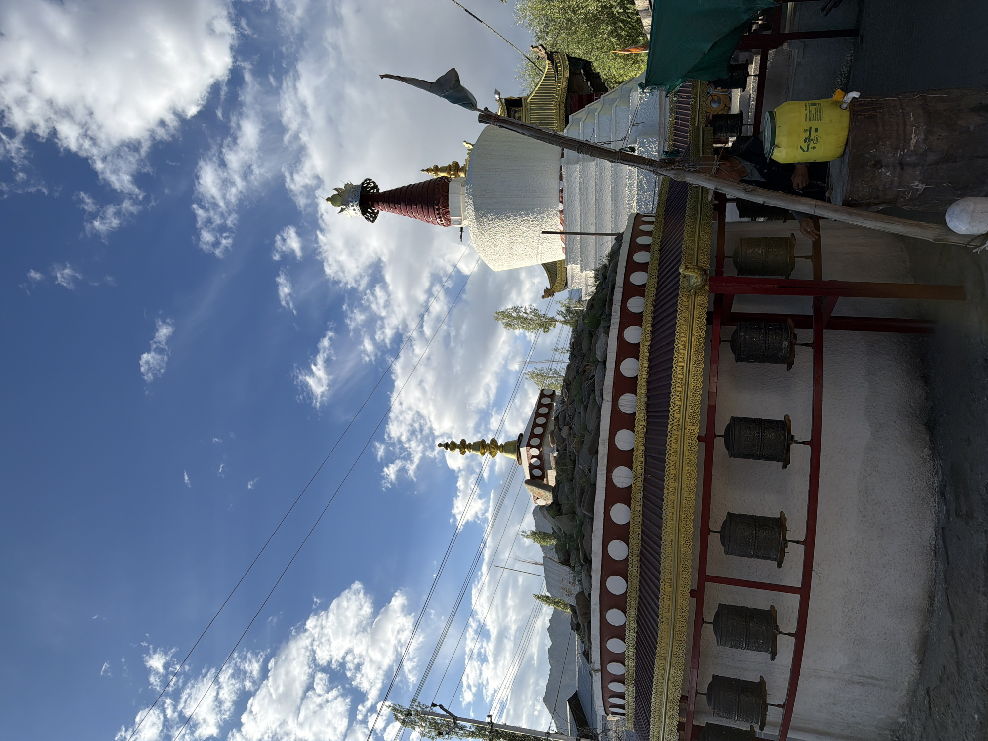 The image depicts a Buddhist stupa with a red spire and white base, situated in the city of Leh, India. In the foreground, a circular structure with a red and gold roof features several prayer wheels mounted on its exterior. The sky above is blue with scattered white clouds, and power lines are visible stretching across the frame. A person is partially visible near a yellow barrel to the right of the prayer wheel structure. The overall scene suggests a religious or cultural site in a mountainous region, likely Leh, given the architectural style and the landscape hints in the background.