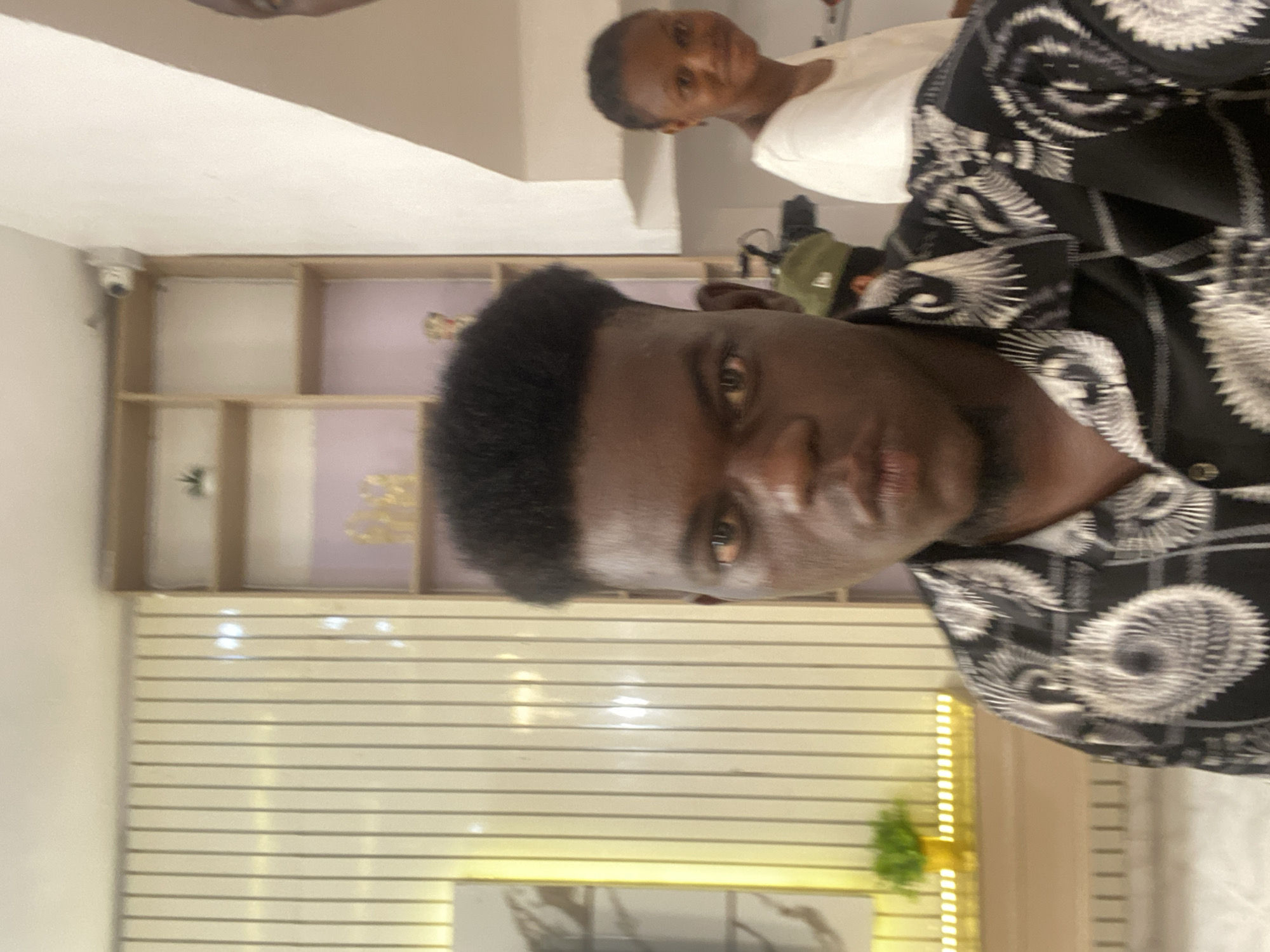 A medium shot shows a man in a black and white patterned shirt. Slightly behind him and out of focus is another person in a white shirt. A security camera is mounted on the wall near a shelf unit with gold accents. A row of horizontal blinds and decorative lighting are visible in the background. Location is Unknown, Unknown.