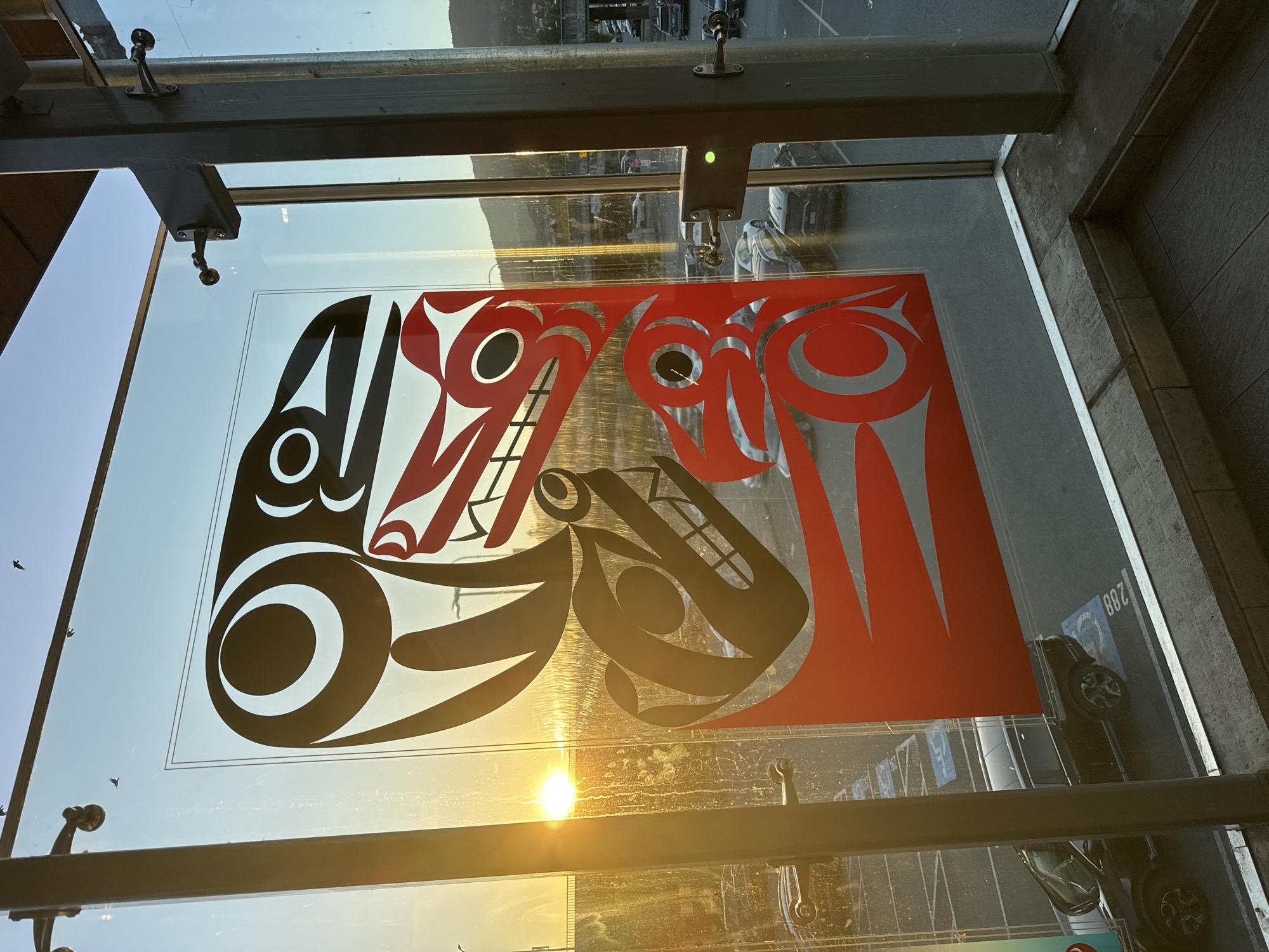 The image shows a large glass panel with a red and black Indigenous design. The sun is setting behind a mountainous landscape, casting a warm glow. In the background, a parking lot filled with cars is visible, along with a bridge and some buildings. Several birds are flying in the sky. The glass panel is mounted on a metal frame structure, with some metal fixtures attached to it. This artwork is located at the Coquitlam Airport, also known as the Boundary Bay Airport, in Coquitlam, Canada.