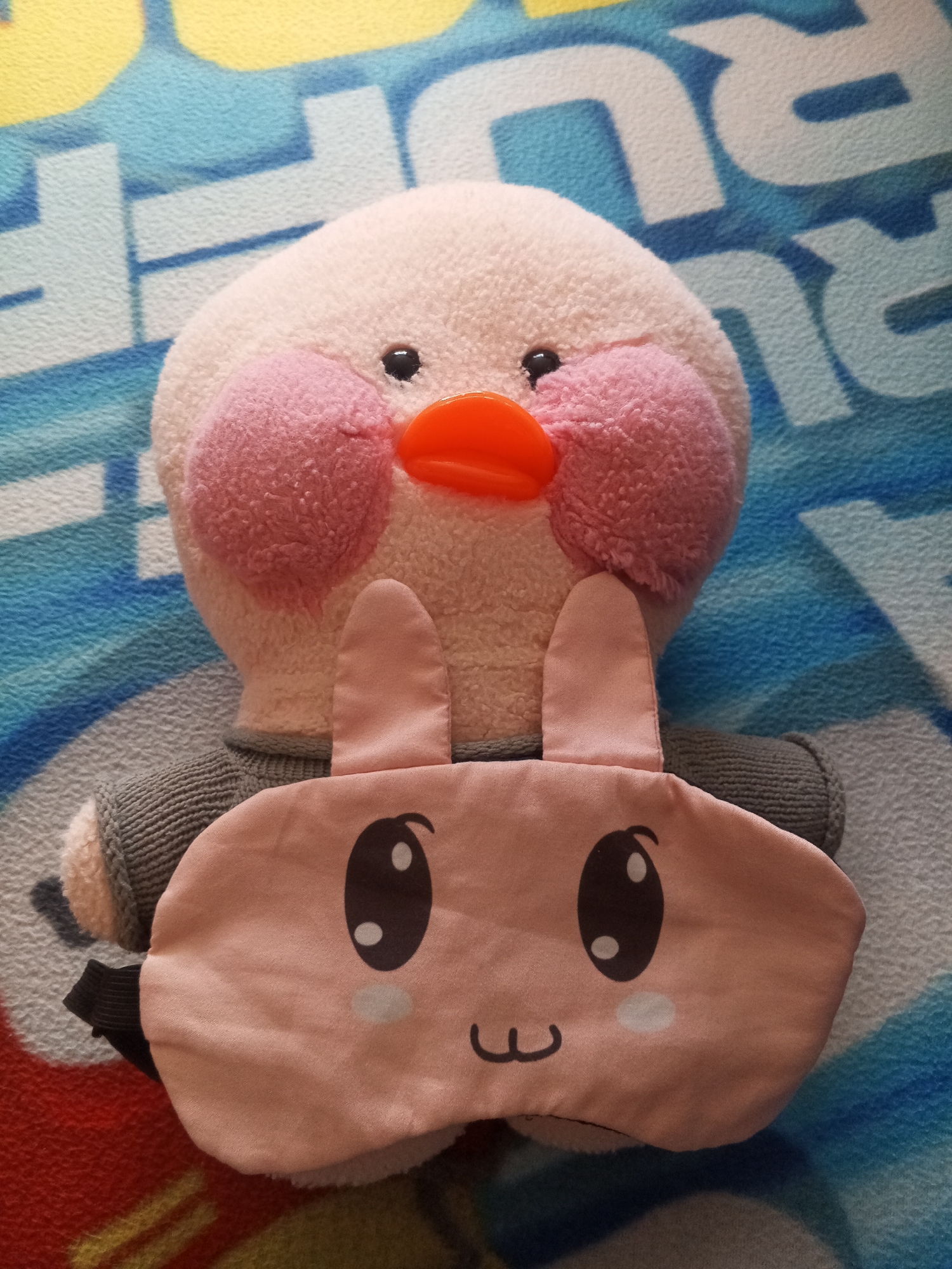 A close-up, top-down shot shows a plush toy duck wearing a pink bunny-themed sleep mask. The duck toy is pale pink with bright orange beak and pink rosy cheeks. The sleep mask is light pink with black outlines of large eyes and a small mouth, and features two upright bunny ears. The toys are resting on a patterned surface, possibly a blanket, with blue and white abstract shapes. The scene is illuminated from above. There is no indication of the location being in Novopokrovskaya, Russia.
