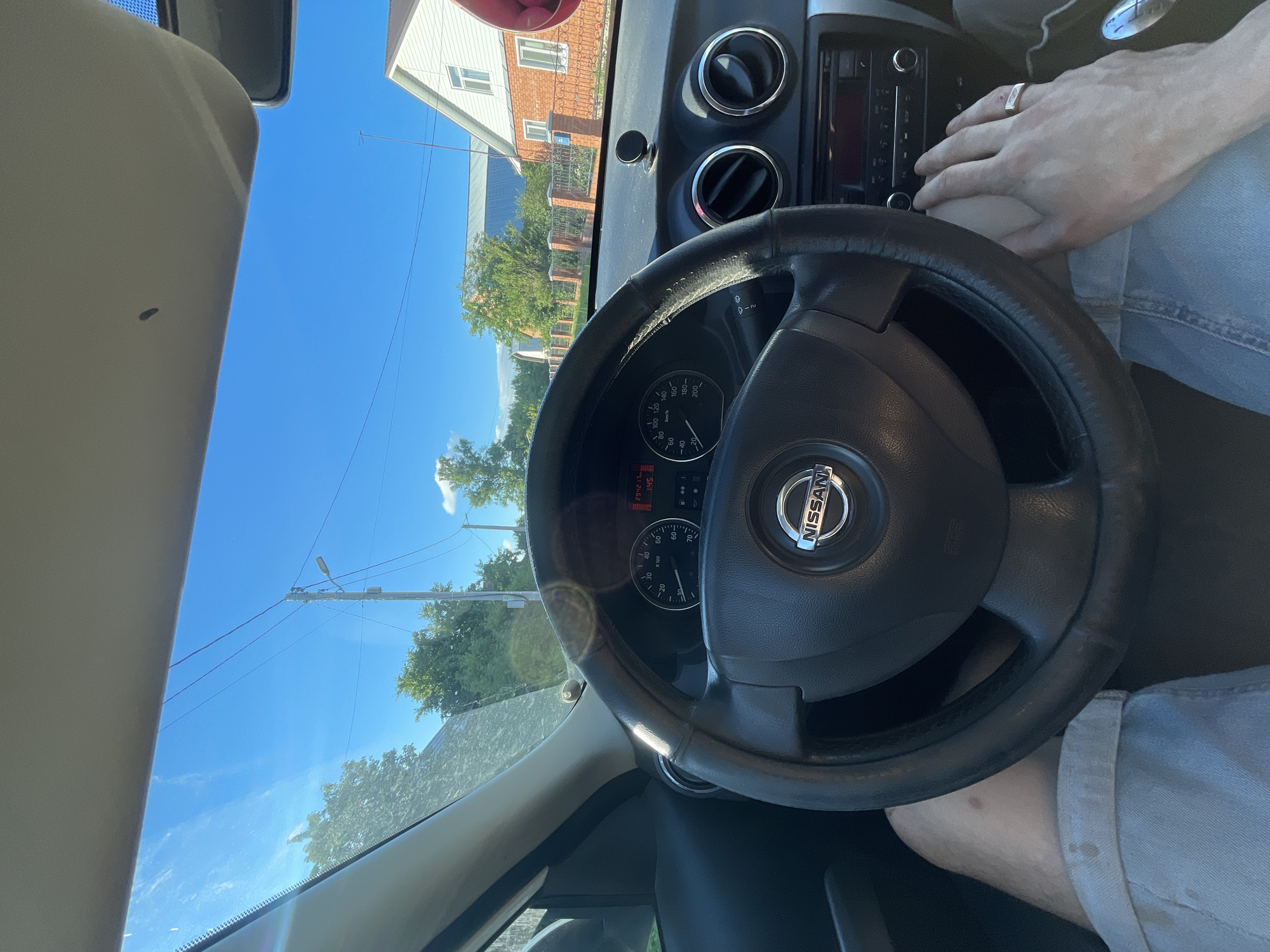 The image shows a first-person perspective from inside a Nissan vehicle, looking out through the windshield. The driver's hands are visible, one resting on the steering wheel and the other reaching towards the car's radio. The dashboard displays a digital readout and the speedometer shows a speed of 0 km/h. Outside, a clear blue sky with sparse clouds is visible, along with trees and houses, suggesting a suburban or rural setting. The scene appears to be in the city of Laishevo, Russia, indicated by the distinct architectural style of the houses and the general ambiance. The lighting suggests it is daytime.