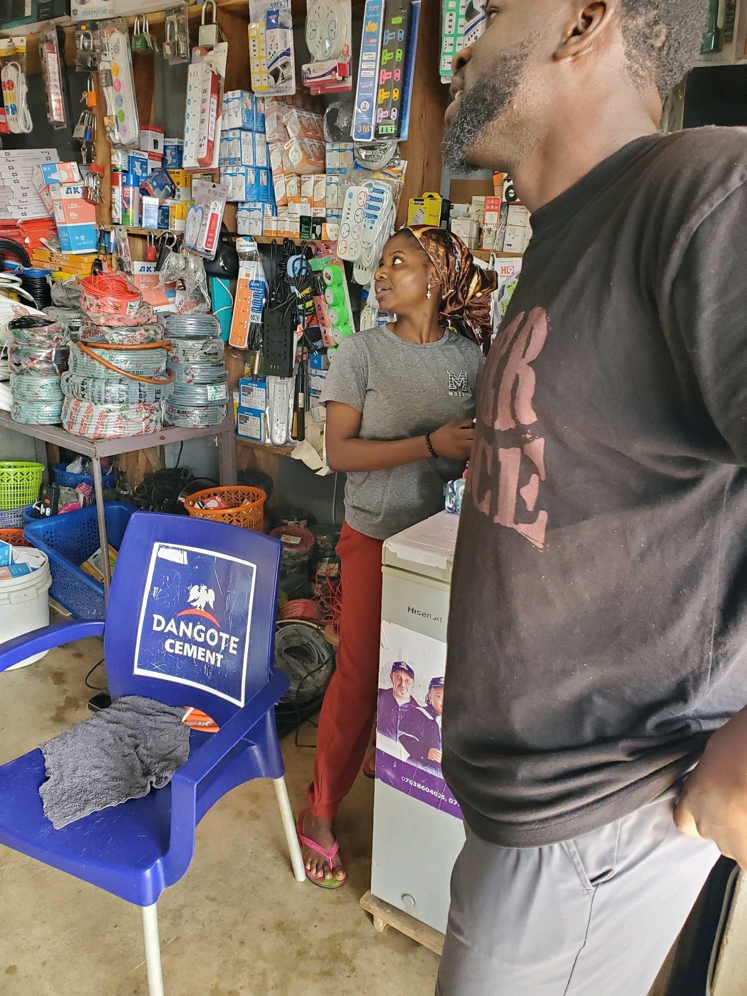 In Gura Top, Nigeria, a man and a woman are standing in a cluttered hardware store. The man, with a beard and wearing a black t-shirt and grey pants, is seen from the chest down, looking to his right. The woman, wearing a grey t-shirt and maroon pants, is looking to her left, engaged in conversation with the man. The store is filled with various electrical supplies, including extension cords, power strips, light bulbs, and wiring, all organized on shelves and racks. A blue chair with the "Dangote Cement" logo is visible in the foreground, with a grey cloth draped over it. A white refrigerator with "Hisense" branding and posters featuring people is located behind the woman. The environment suggests a typical retail space in a Nigerian city, characterized by its dense display of goods and the presence of customers interacting with a salesperson or fellow shopper.