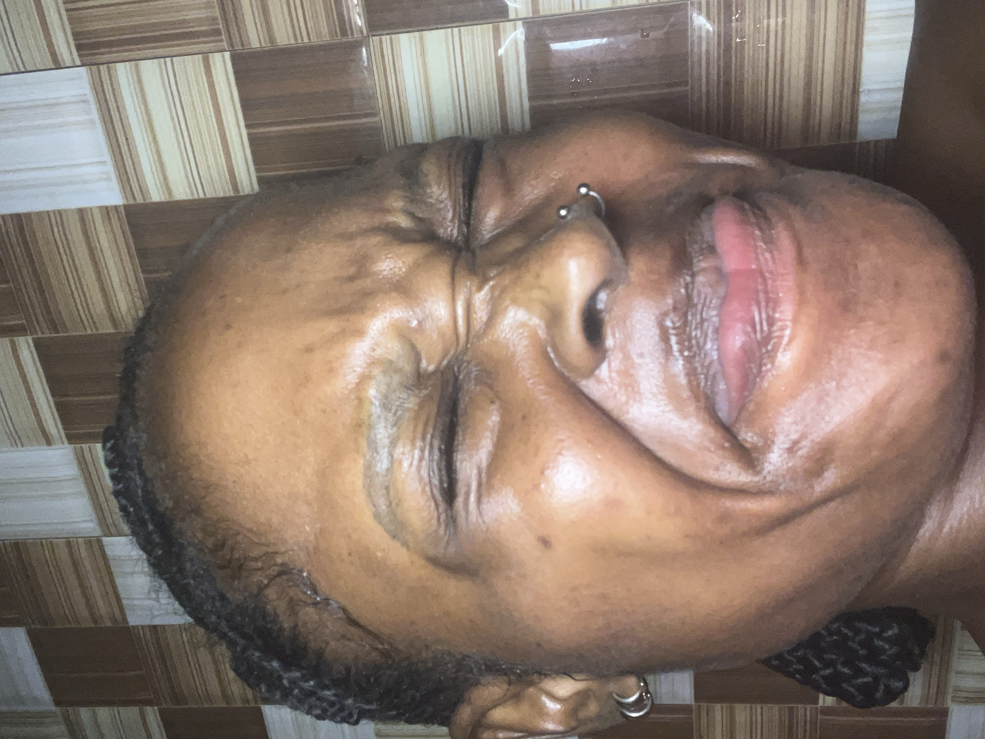 A close-up, low-angle, head-and-shoulders shot shows a Black woman with her eyes squeezed shut and her face contorted as if in discomfort or disgust. Her mouth is slightly open, and her tongue is visible. She has braids woven into her hair, a nose ring, and earrings. The background is a tiled wall. The image was taken in a bathroom in Ado, Nigeria.