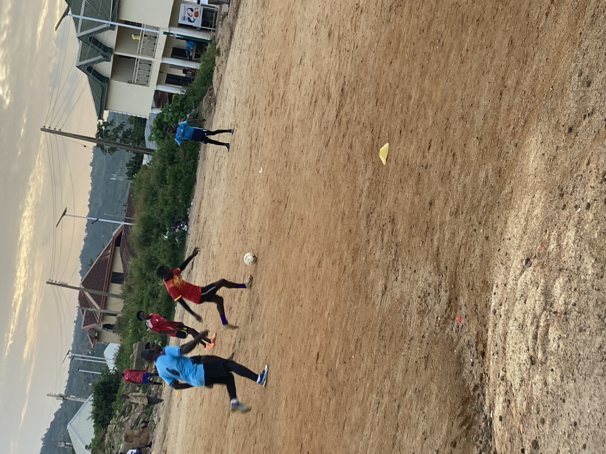 A group of young men are playing soccer on a dirt field in an urban area of Jos, Nigeria. In the foreground, a young man in a blue jersey and black shorts is running with the ball at his feet. To his right, another young man in a red and yellow jersey is also running, with his eyes on the ball. In the background, several other young men in similar athletic attire are visible, some watching the game and others participating. The field is uneven and dusty, with scattered rocks and debris. Behind the field, there are buildings, including a multi-story building with a green roof, and a hill covered in vegetation. The sky is overcast with soft clouds. The image captures a moment of casual athletic activity in a developing urban landscape.