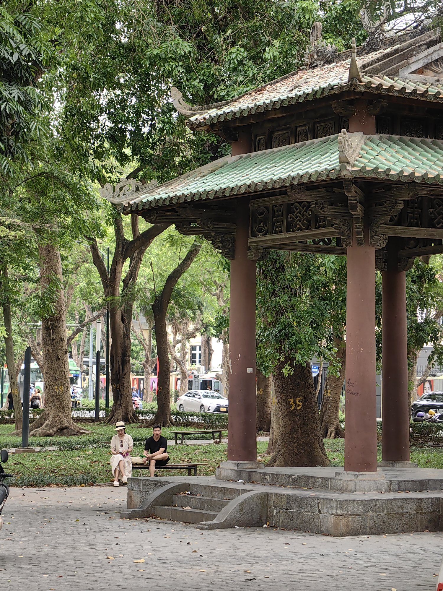 The image shows a pagoda-like structure in a park setting. The structure has a green tile roof and is supported by brown pillars. Two people are sitting on a bench nearby. The park appears to be located in a city, with cars and other vehicles visible in the background. The location is likely in Hà Nội, Vietnam, based on the architectural style of the pagoda. A large tree with the number 35 painted on its trunk is visible near the pagoda. The area around the pagoda is paved with stone.