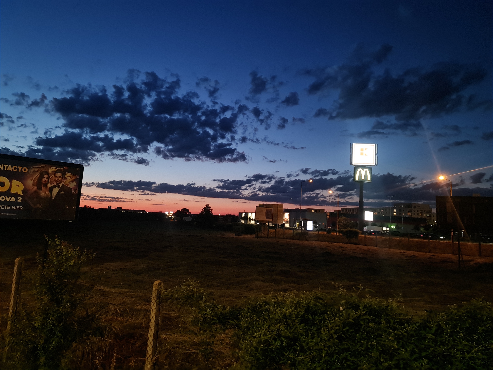 This image depicts an outdoor evening scene in the city of Unknown, Slovakia, characterized by the transition from dusk to night. The sky displays a deep blue overhead, with scattered dark, cumulus-like clouds. Towards the horizon, particularly on the left, the sky transitions through hues of purple and vibrant orange-red, indicative of a sunset.
In the foreground, an unkempt grassy field occupies the lower half of the frame. Sparse green bushes and taller weeds are visible, with two faded wooden fence posts situated in the lower left.
The midground features a large, illuminated billboard on the left. The visible text fragments include "NTACTO," "OR," "NOVA 2," and "ETE HIER." The advertisement prominently features two individuals: a man in a dark suit and a woman in a formal dress.
Towards the right midground and background, a commercial area is visible. A tall pole supports two illuminated signs: a square white sign with a stylized "K" logo on top, and a rectangular white sign with the golden "M" logo of McDonald's beneath it. Several other illuminated buildings and smaller signs are present. Streetlights are active, casting an orange glow. A road or parking area with indistinct vehicles is implied behind the commercial establishments. Distant buildings are silhouetted against the fading light of the sunset. No human figures are discernible in the scene itself, apart from those depicted on the billboard.