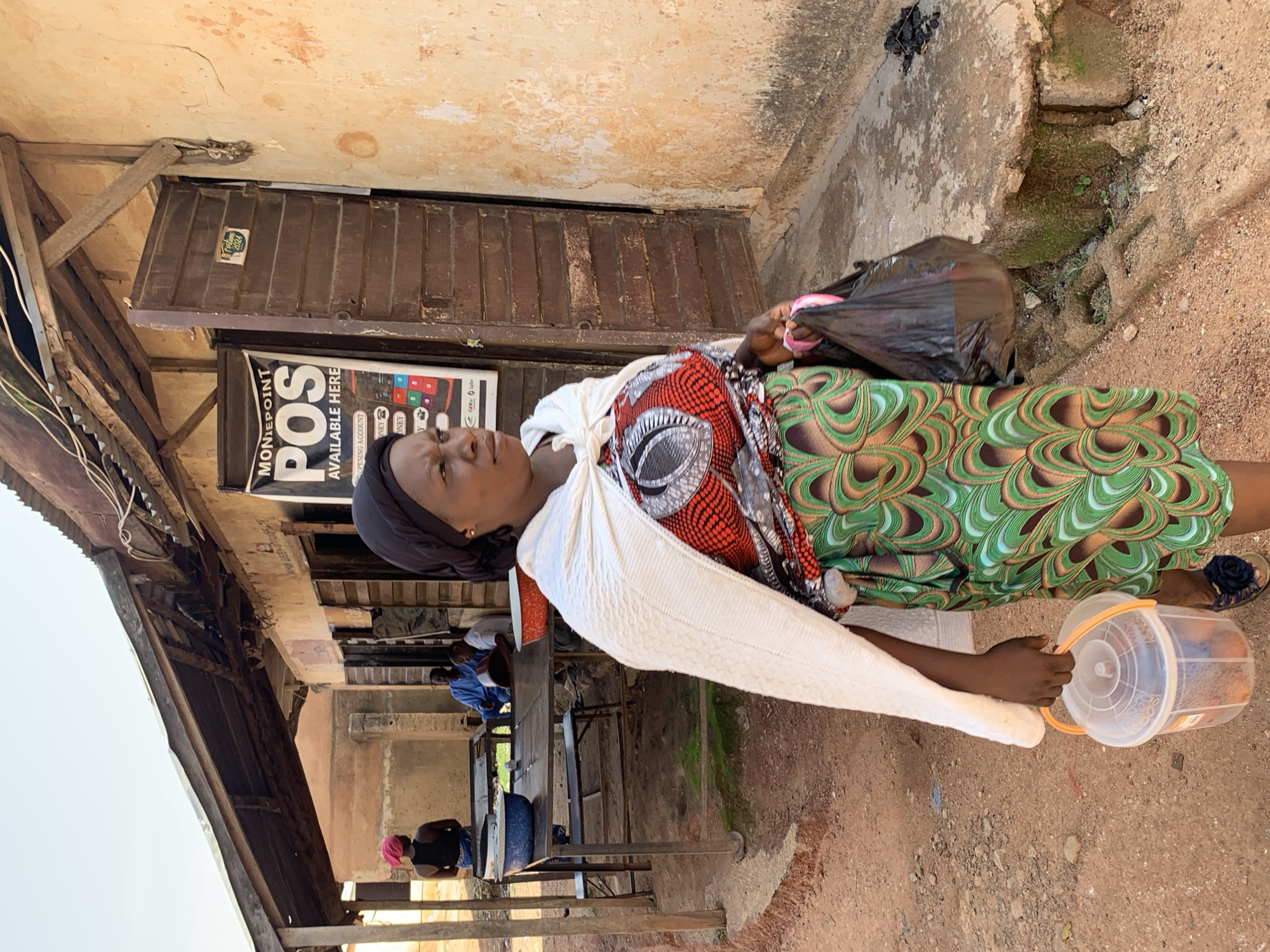 This is a portrait of a Black woman standing outdoors in a sandy, dusty area, possibly a market or a street in Jos, Nigeria. She is wearing a black headscarf, a white shawl draped over her shoulders, and a vibrantly patterned dress with green, brown, and white swirls. She is holding a black plastic bag in her right hand and a clear plastic bucket with an orange lid in her left hand. The bucket appears to contain several orange objects, possibly fruits or vegetables.
Behind her, a building with a corrugated metal roof and wooden supports is visible. A sign on the building reads "MONIEPOINT POS AVAILABLE HERE" with colorful icons of credit cards and mobile phones. The building's facade is weathered, with cracked paint and a wooden door. In the background, other individuals are present, some sitting at tables and others standing. One person in the background wears a pink headscarf.
The scene appears to be set during the daytime. The lighting suggests it might be a warm, possibly sunny day. The overall impression is one of everyday life and commerce in a busy, perhaps informal, commercial area. The woman's expression is difficult to discern clearly, but she seems focused on the items she is carrying. The presence of the "Moniepoint POS" sign suggests a location where financial transactions might occur, indicating a commercial establishment.