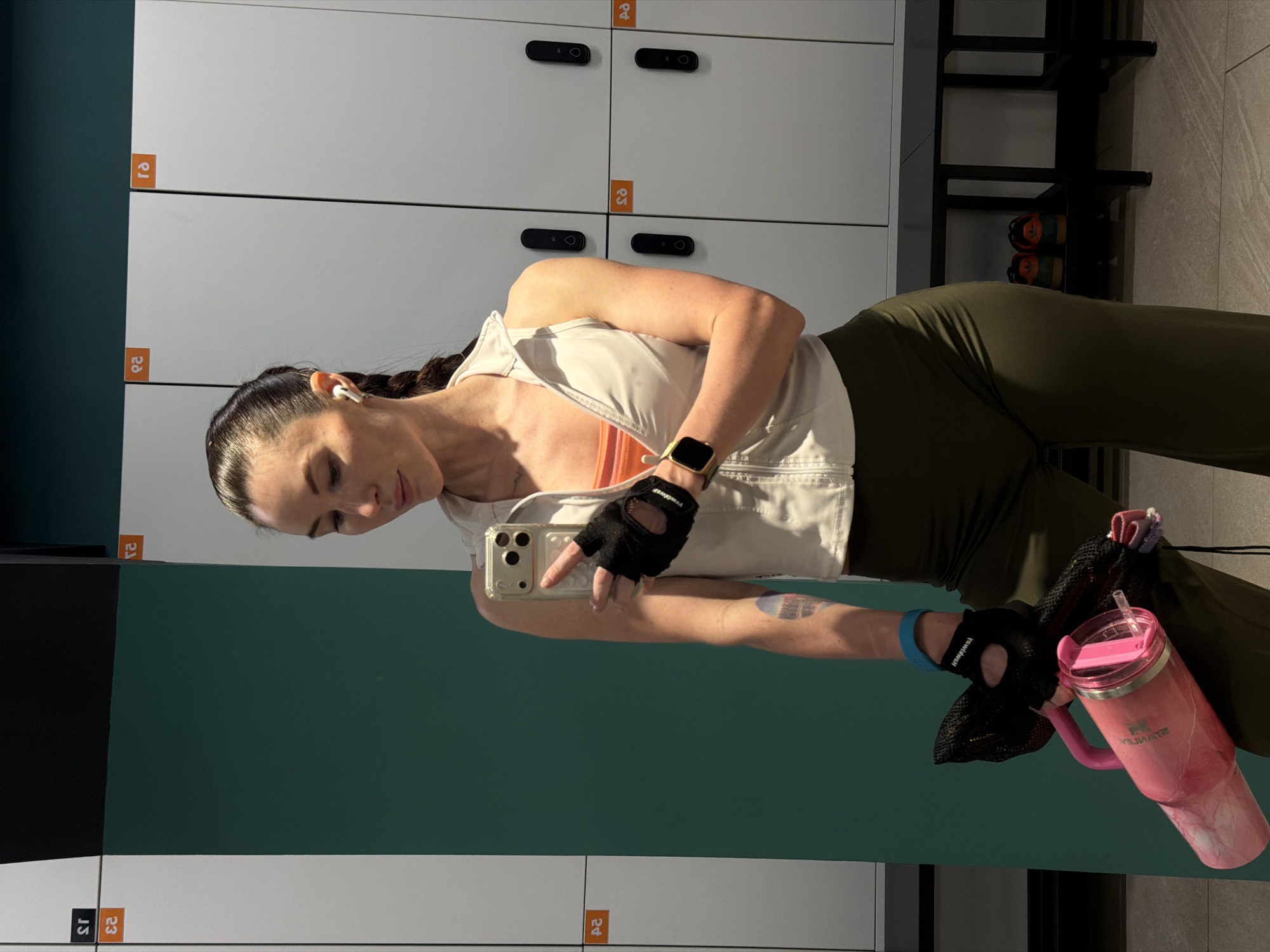 A woman is taking a selfie in front of lockers. She is wearing fitness attire: olive green leggings, an orange top, a white vest, and black fingerless gloves. She has dark hair pulled back in a braid and is wearing wireless earbuds. Her phone is in her left hand and her right hand is extended to the screen, she is wearing a smartwatch on the right arm. She is holding a pink Stanley brand tumbler inside of a black mesh bag. The lockers are light gray with black handles and orange number labels. The lockers are stacked in rows. A dark teal wall section is to the left of the lockers. A black shoe rack with athletic shoes on the shelves is visible in the background. The scene appears to be in a gym locker room. It is impossible to verify the location is the city of Perm, Russia.