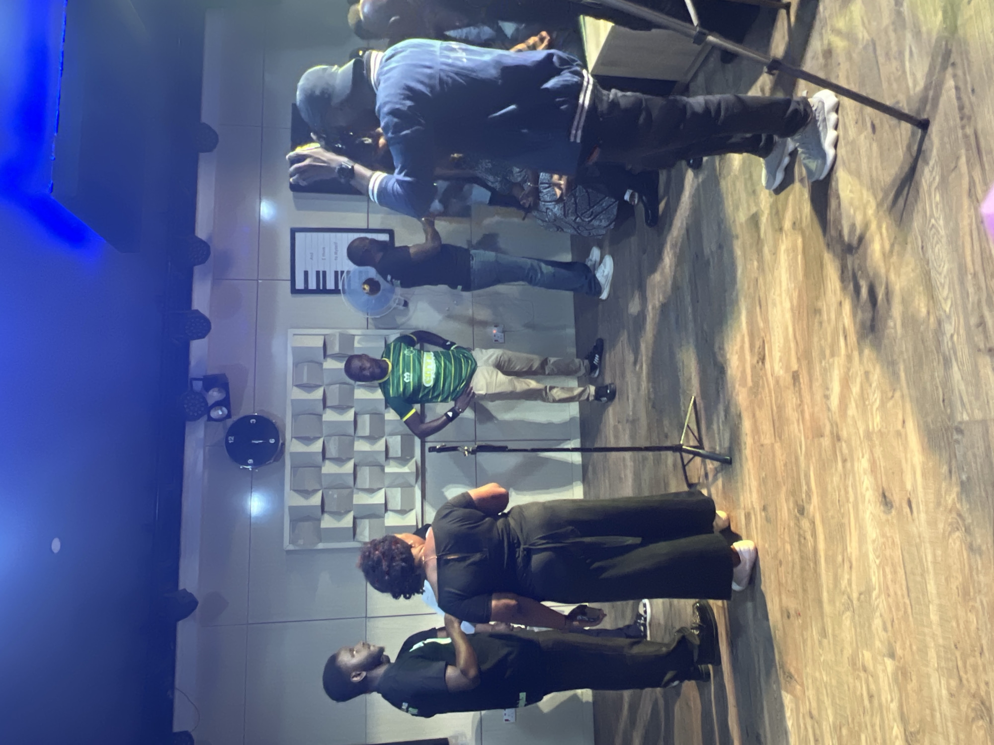 A group of individuals are gathered in what appears to be a studio space, likely in Jos, Nigeria. The room has a wooden floor and a wall decorated with a geometric pattern. Lighting fixtures are visible on the ceiling. In the foreground, a woman in a dark dress stands facing away from the camera, while two men in black t-shirts are to her left. To the right, a man in a green and white striped shirt stands, and another man in a blue jacket and a cap is facing the woman, appearing to take a picture with a camera. Several other individuals are partially visible in the background, some seated. A clock is mounted on the wall.