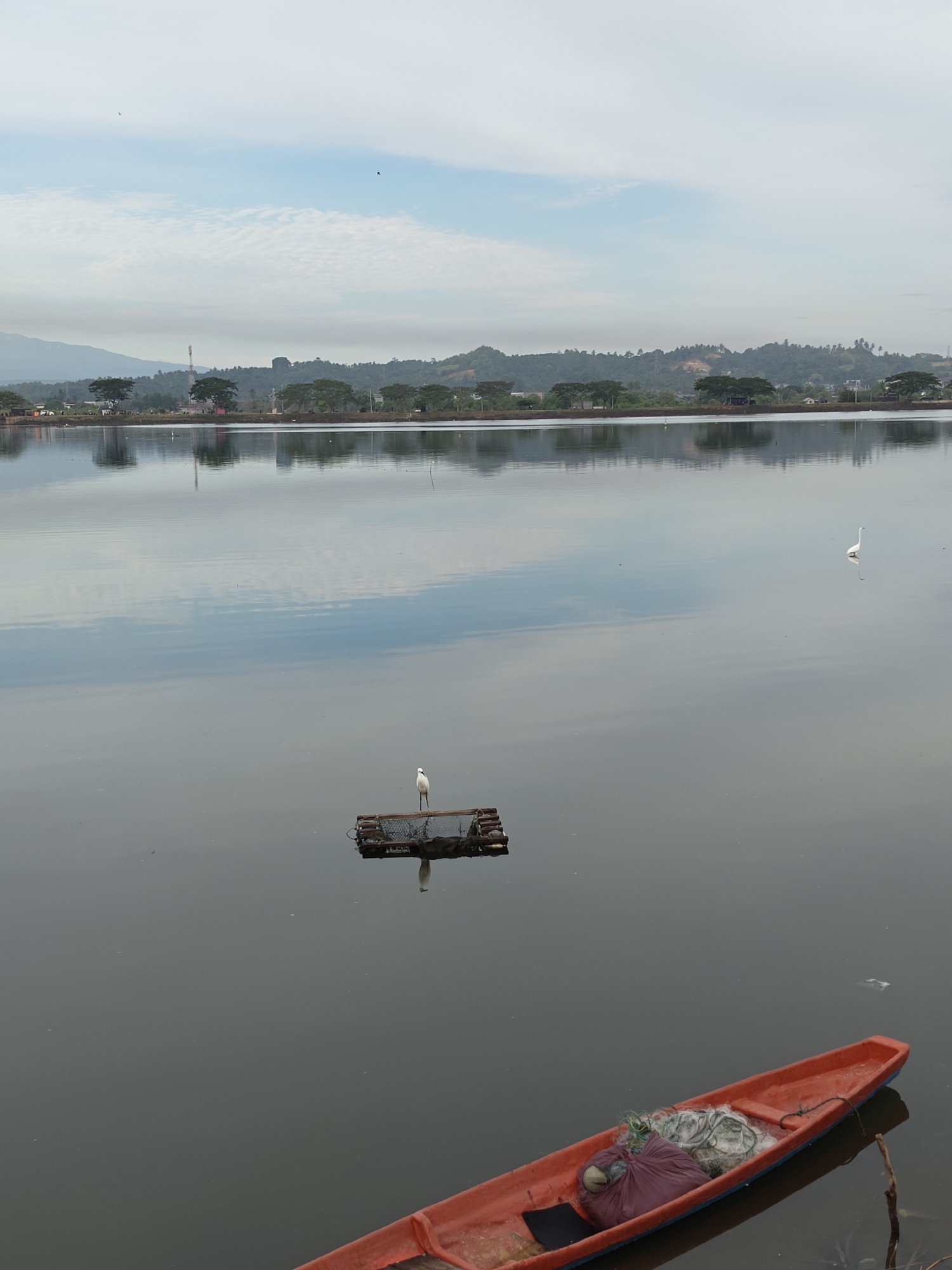 The image depicts a serene body of water, likely a lake or a calm bay, with a reflective surface that mirrors the cloudy sky and the distant treeline. In the foreground, a bright orange canoe rests on the water, filled with bundles of fishing nets and a large purple bag. Further out in the water, a small, dark raft floats, occupied by a lone white egret. Another white egret is visible in the distance, to the right of the raft. The background reveals a gentle, rolling landscape covered in lush green trees, interspersed with scattered buildings and what appears to be a telecommunications tower. The overall atmosphere is tranquil and picturesque, suggesting a quiet morning or late afternoon in a natural setting. This scene is likely located near the coastal areas of Lhokseumawe, Indonesia, known for its natural beauty and fishing communities.