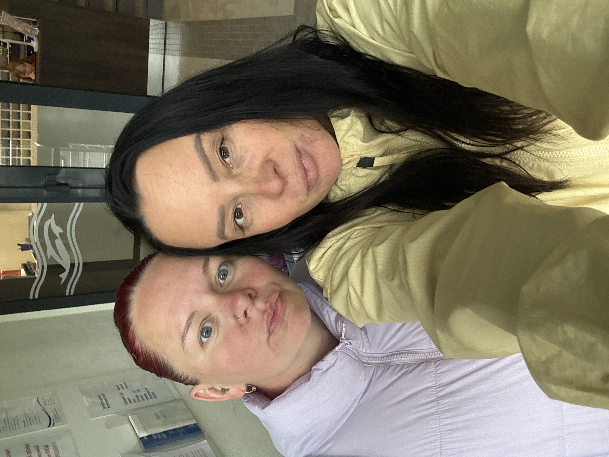 Two women are posing for a selfie indoors. The woman on top has dark hair and is wearing a light yellow jacket. The woman on the bottom has short red hair and is wearing a light purple jacket. The background appears to be an office setting. There are papers and a window behind them. It is difficult to say the location is in Chaikovsky, Russia based on the image provided.