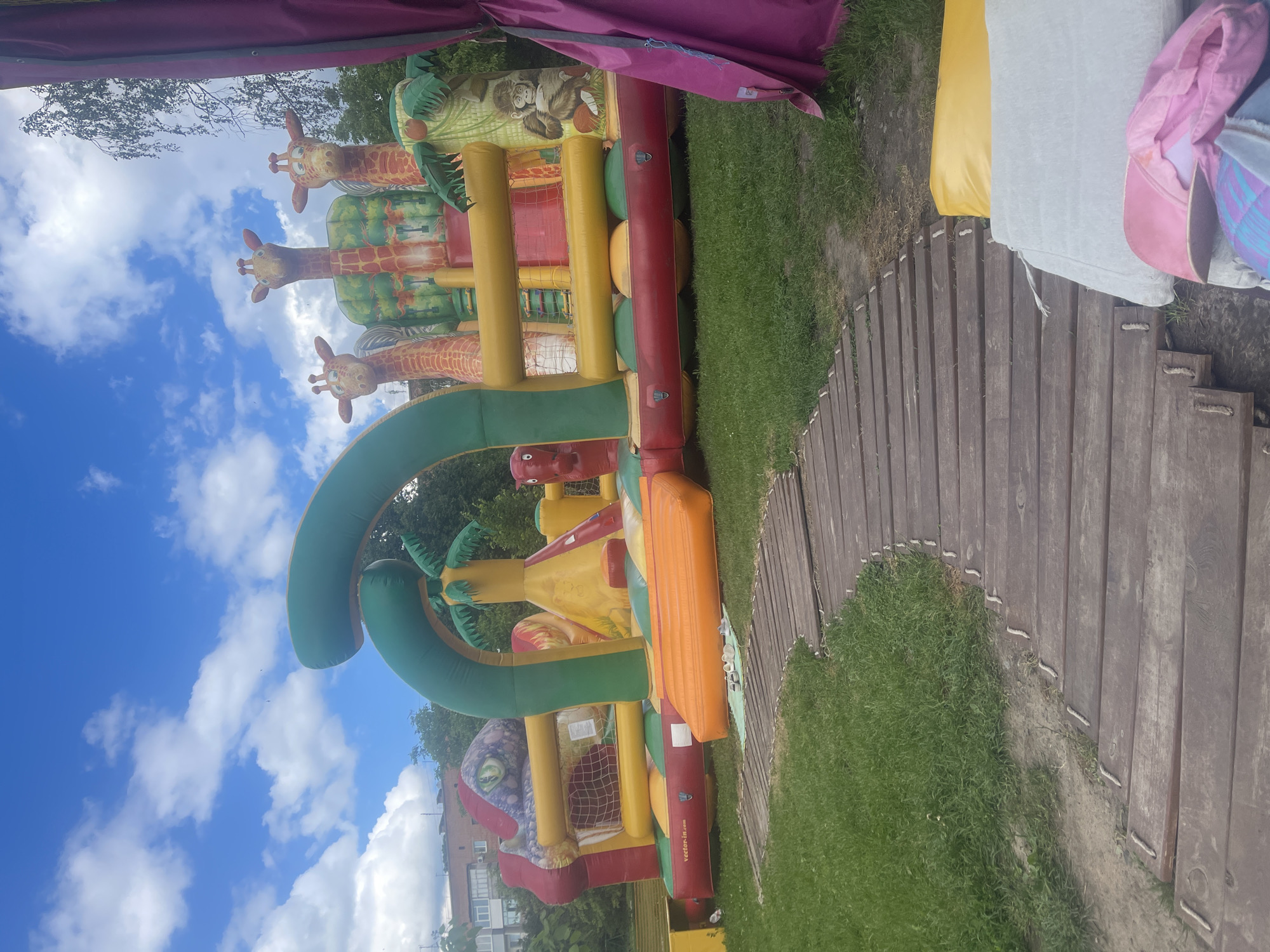 In an outdoor grassy area, a large inflatable playground is set up. The playground has a jungle theme, featuring a dinosaur head, giraffes, a monkey, and palm trees. The structure is colorful, with red, yellow, green, and orange sections. In the foreground, a wooden walkway leads towards the playground, and there are some cushions and a pink bag on the ground next to it. The sky is blue with scattered white clouds, and trees are visible in the background.